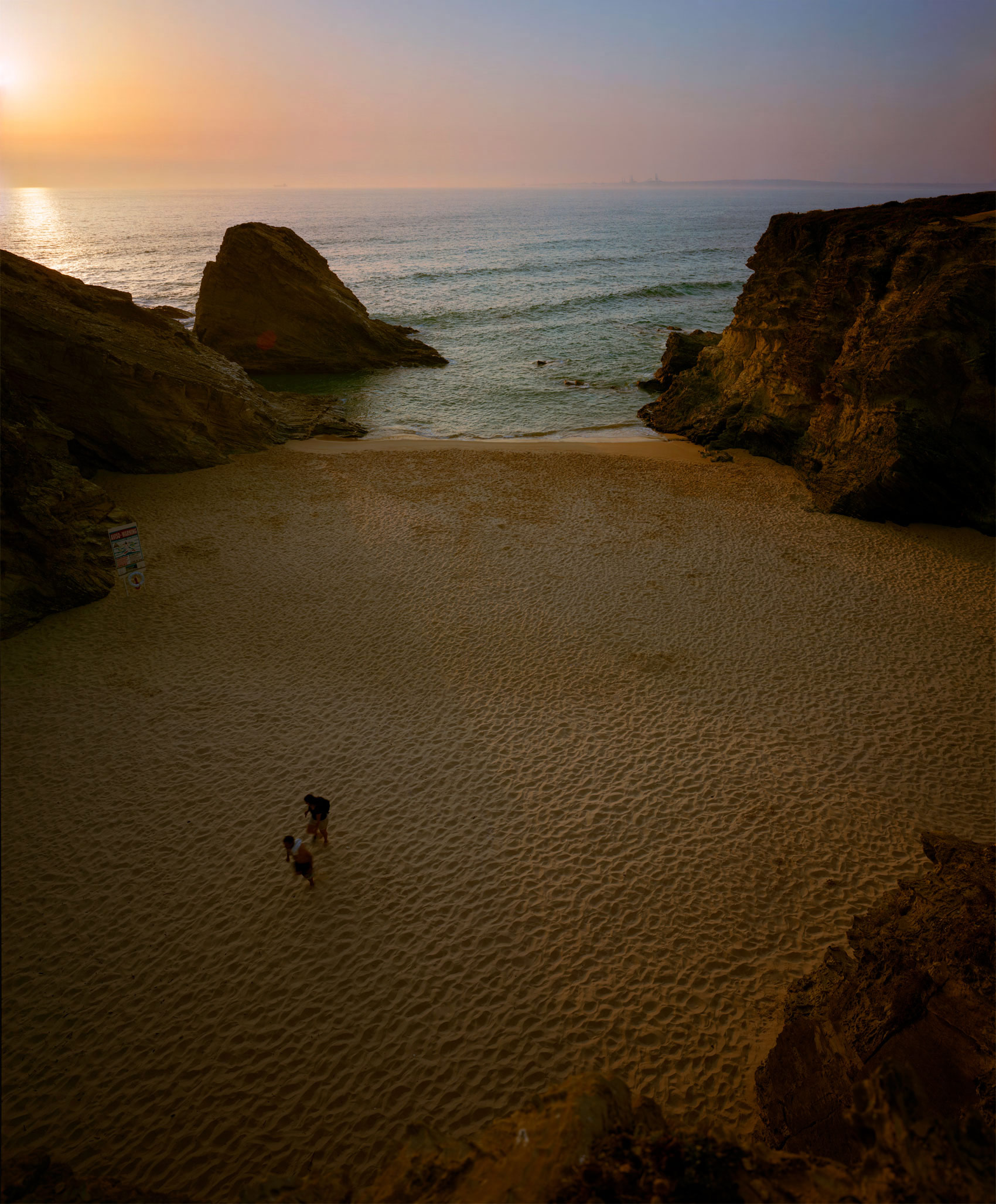 A secluded sandy beach framed by dark rocky outcrops on either side, with a solitary figure walking across the pale sand toward turquoise waters. The sky glows with warm golden and pink tones of sunset or sunrise, creating a luminous reflection on the calm sea horizon.