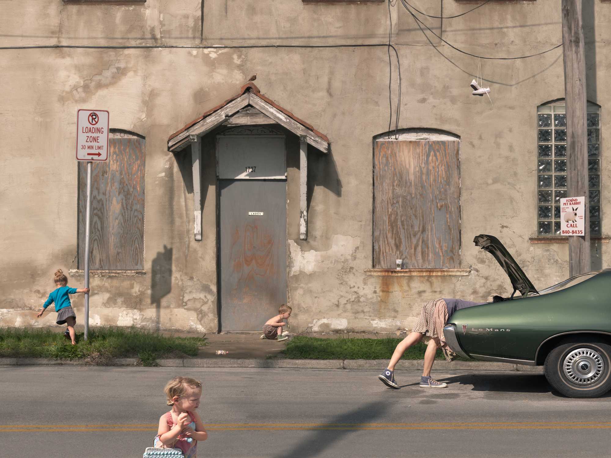 A weathered tan building facade with boarded windows and a central door features children at play on the street below. A young girl in a pink shirt stands in the foreground, while others in bright clothing run and move near a vintage green convertible with its hood open, creating a scene of everyday childhood activity juxtaposed against urban decay.