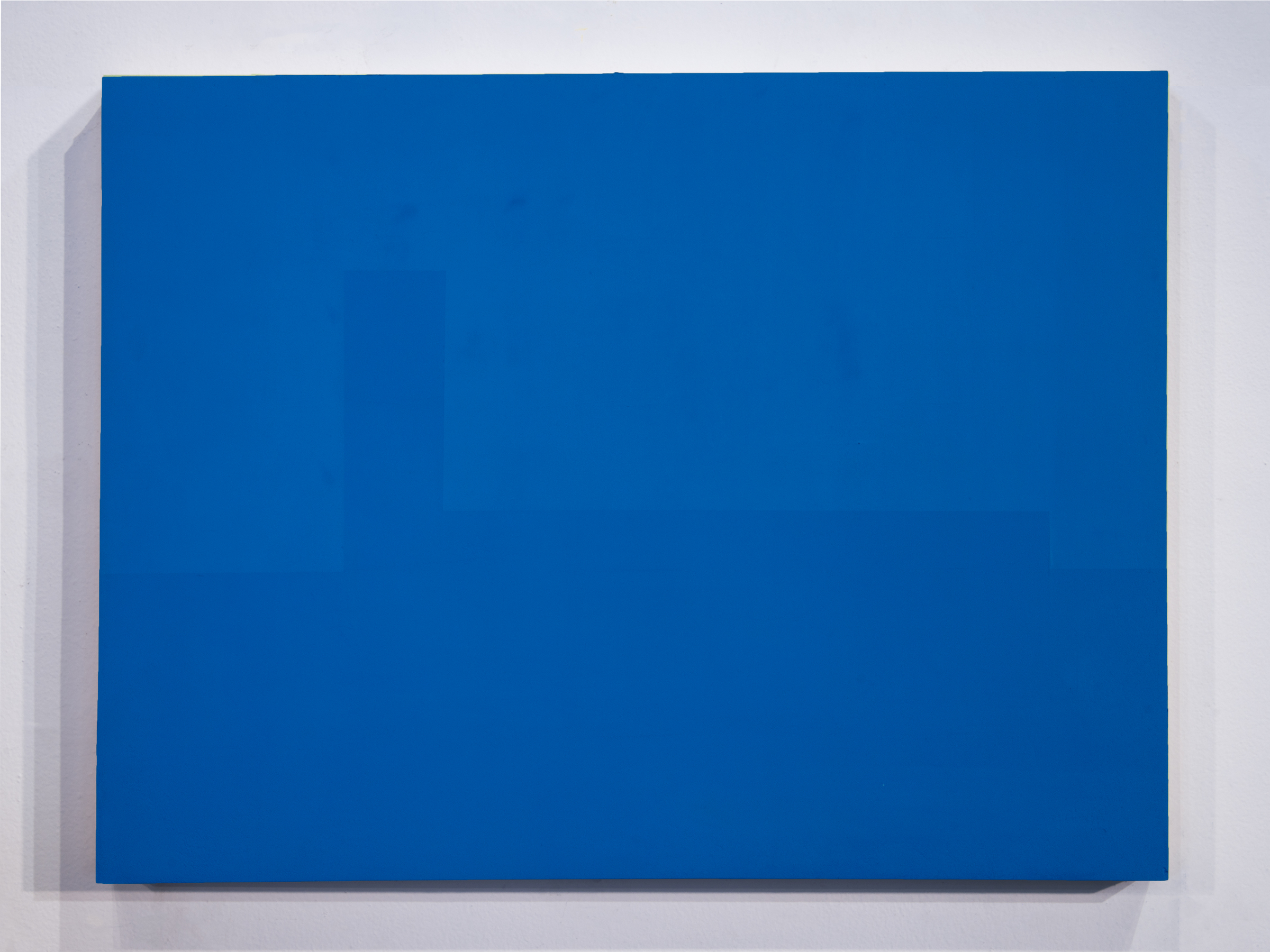 A vibrant blue rectangular panel mounted on a light gray wall, casting a subtle shadow. The monochromatic composition emphasizes the pure saturation of the blue pigment and the work's geometric minimalism.