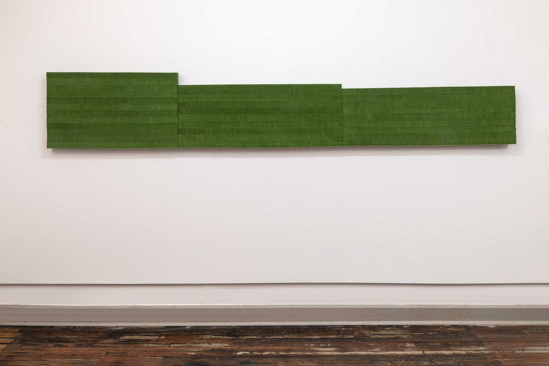 A horizontal green wool felt panel with stepped edges mounted on a white gallery wall above dark wooden flooring. The work consists of three rectangular sections of varying heights arranged in a staggered composition, creating a subtle architectural rhythm across the wall.