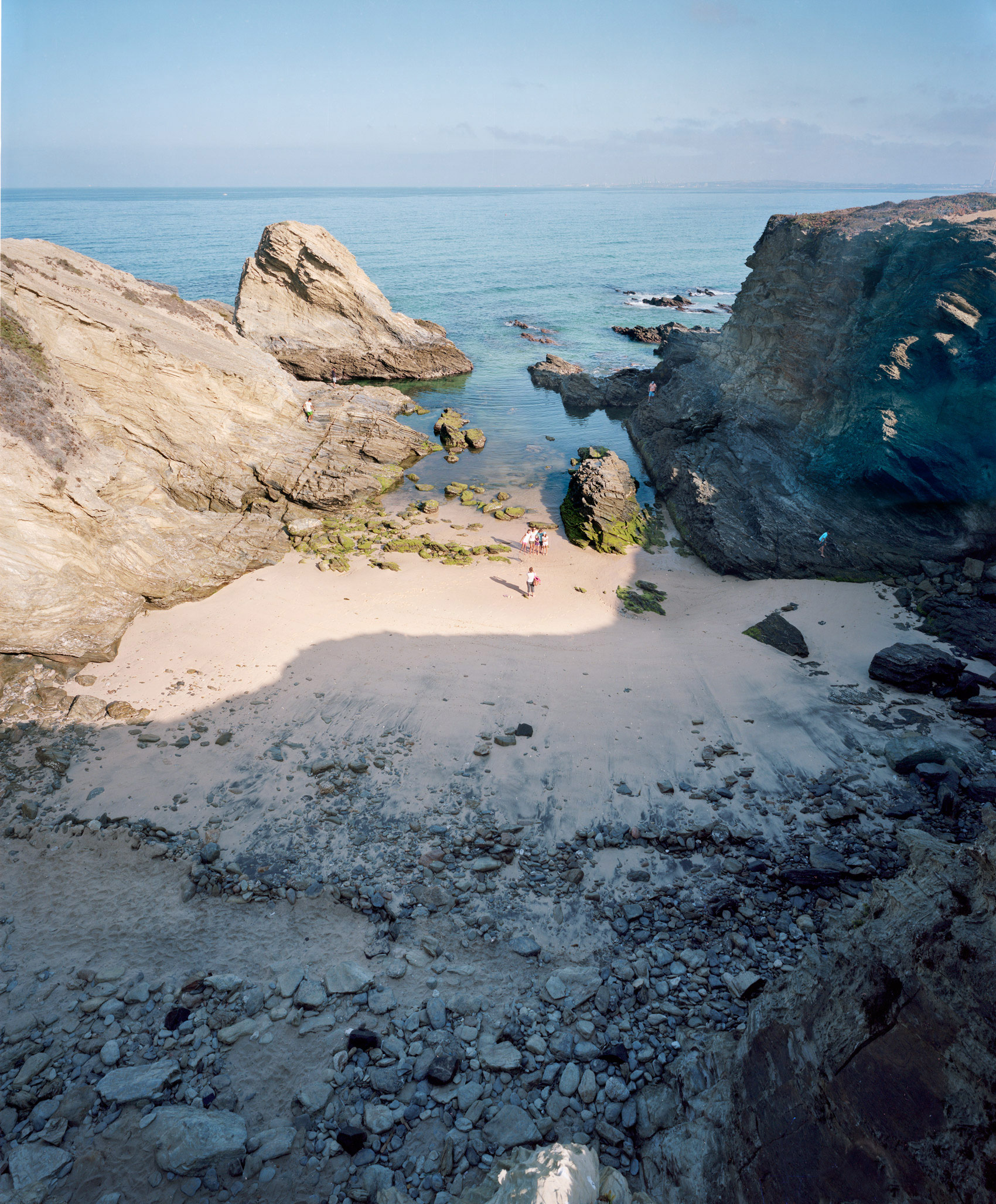 A secluded rocky cove with pale sand nestled between dramatic stone cliffs and outcroppings, the turquoise water beyond dotted with moss-covered rocks. A few visitors in light clothing stand on the beach, dwarfed by the towering geological formations framing the scene under clear sky.