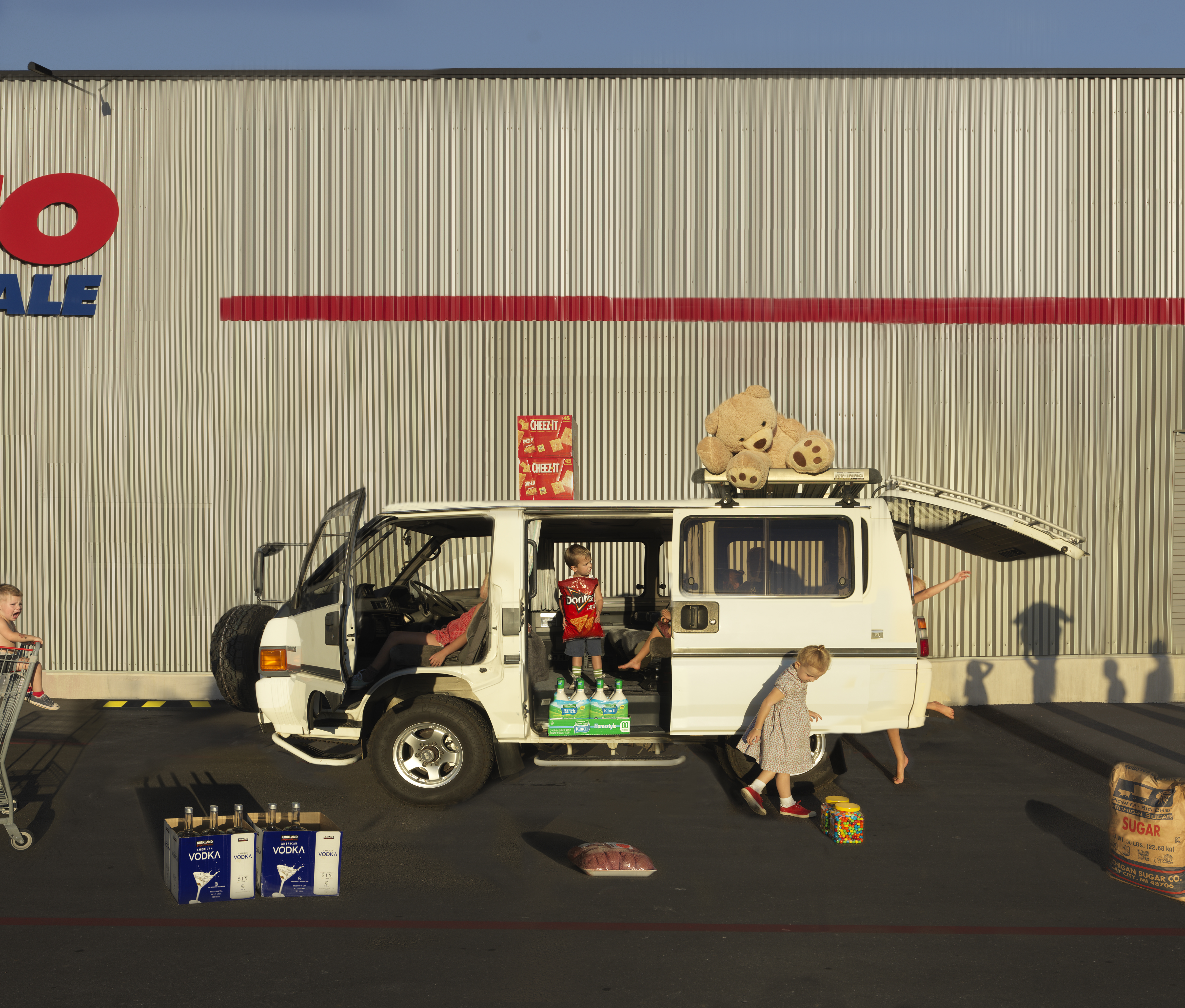 A white minivan loaded with consumer goods and a large teddy bear sits parked in front of an industrial corrugated metal warehouse backdrop bearing a red corporate logo. Children and adults are arranged around the vehicle amid scattered products including vodka bottles, crackers, and a bag of sugar, creating a surreal domestic scene that subverts typical consumer culture imagery through deadpan composition and saturated color.