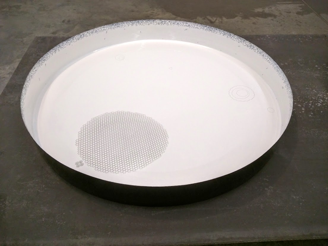 A large circular white porcelain enamel bowl with a textured rim sits on a dark concrete floor. At its center, a perforated metal disk is surrounded by faint algorithmic line drawings etched into the white surface.