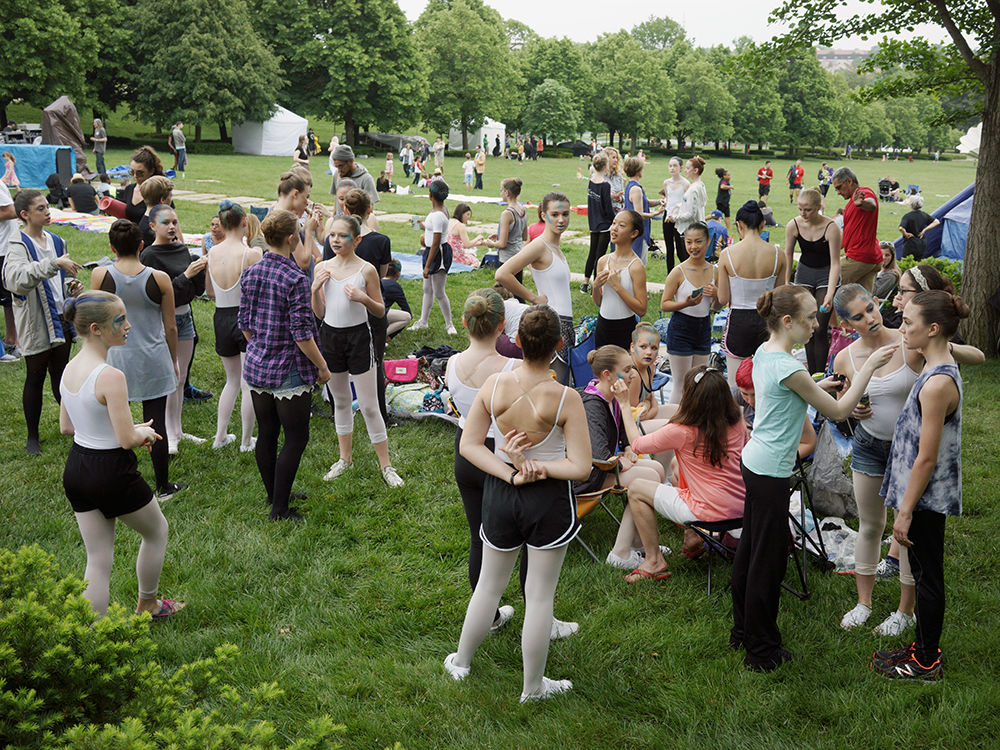 A large gathering of people, predominantly young women and girls in athletic wear, assembled on a sprawling green lawn dotted with trees. The crowd stands in loose clusters amid blankets and folding chairs, with a white tent visible in the distance, suggesting an organized outdoor event or festival in a park setting.
