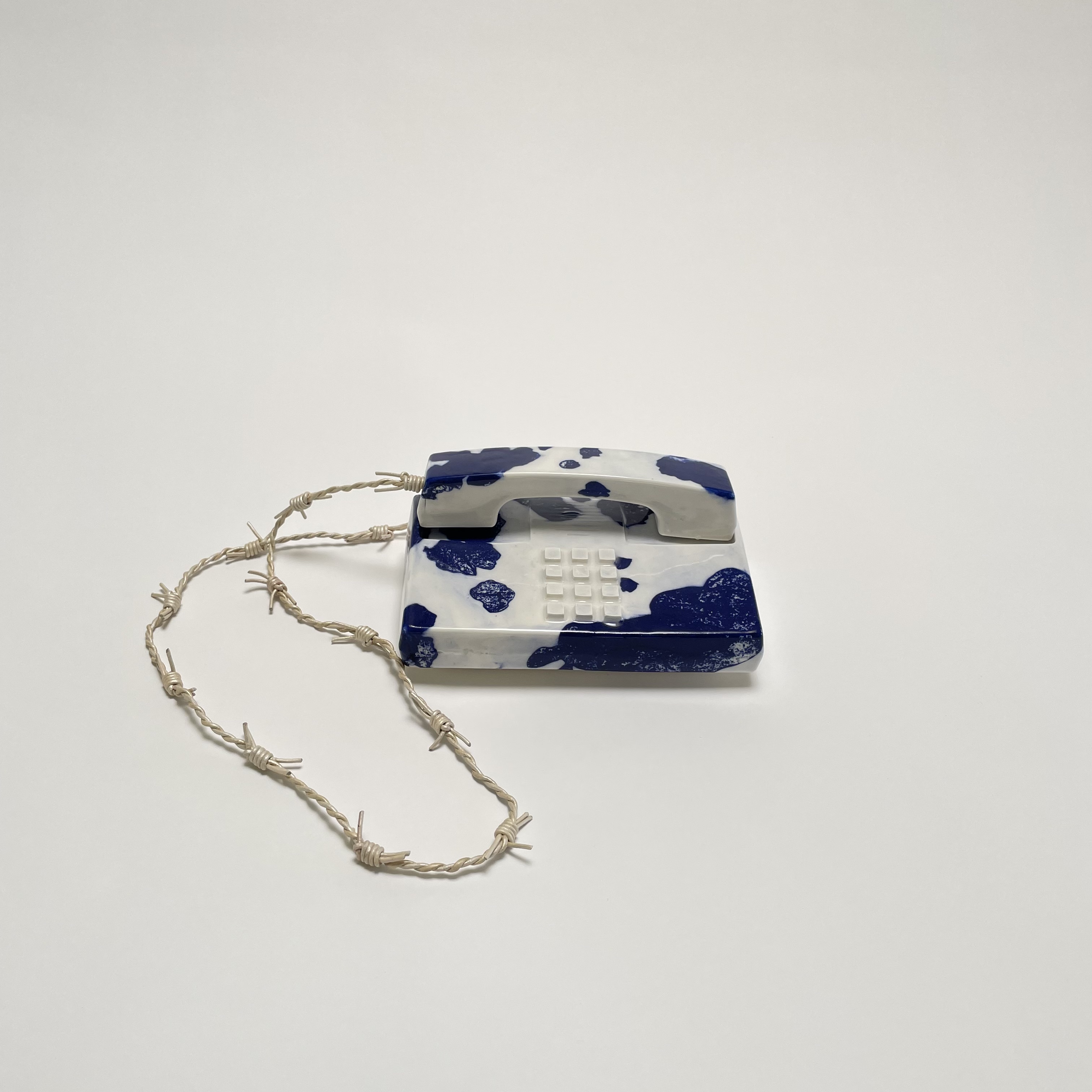 A rectangular porcelain tag with cobalt blue and white cow print pattern, featuring a raised grid of small white dots in the center, suspended from a knotted natural fiber cord.