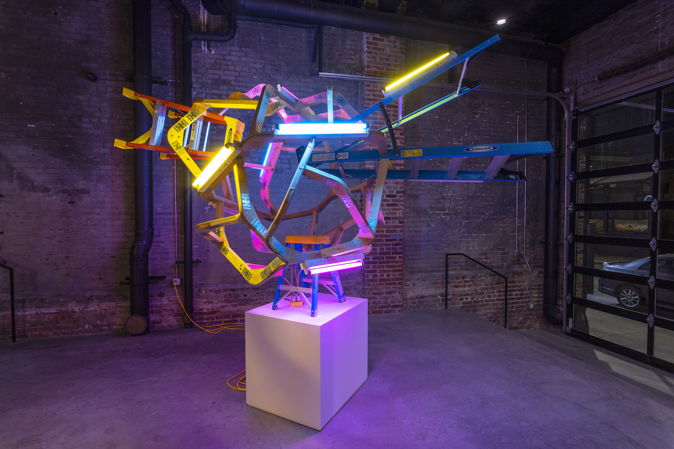 Neon and fluorescent lights in yellow, pink, blue, and green intertwine across repurposed step ladders mounted on a white cubic pedestal, creating an energetic abstract sculpture against exposed brick walls in an industrial gallery space.