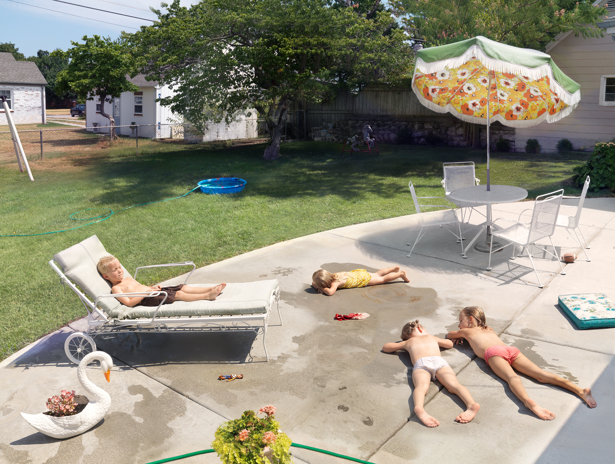 A suburban backyard scene showing multiple figures relaxing on a concrete patio: an adult on a lounge chair, children lying on the ground, and another figure in the distance. A yellow and green striped umbrella shades a table with chairs, while a blue trampoline and various toys are visible on the grass beyond. White houses and trees frame the composition under bright daylight.