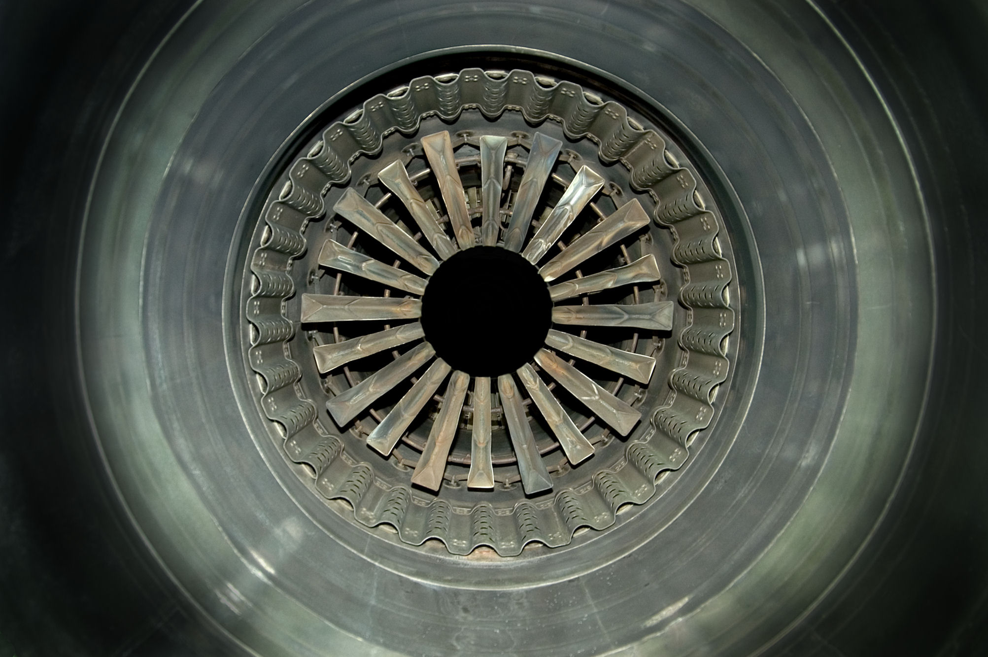 Frontal view of a jet engine intake, showing concentric circles of metallic blades and ducts radiating from a central black aperture. The photograph captures the geometric precision and industrial symmetry of the engine's interior in cool gray and silver tones.