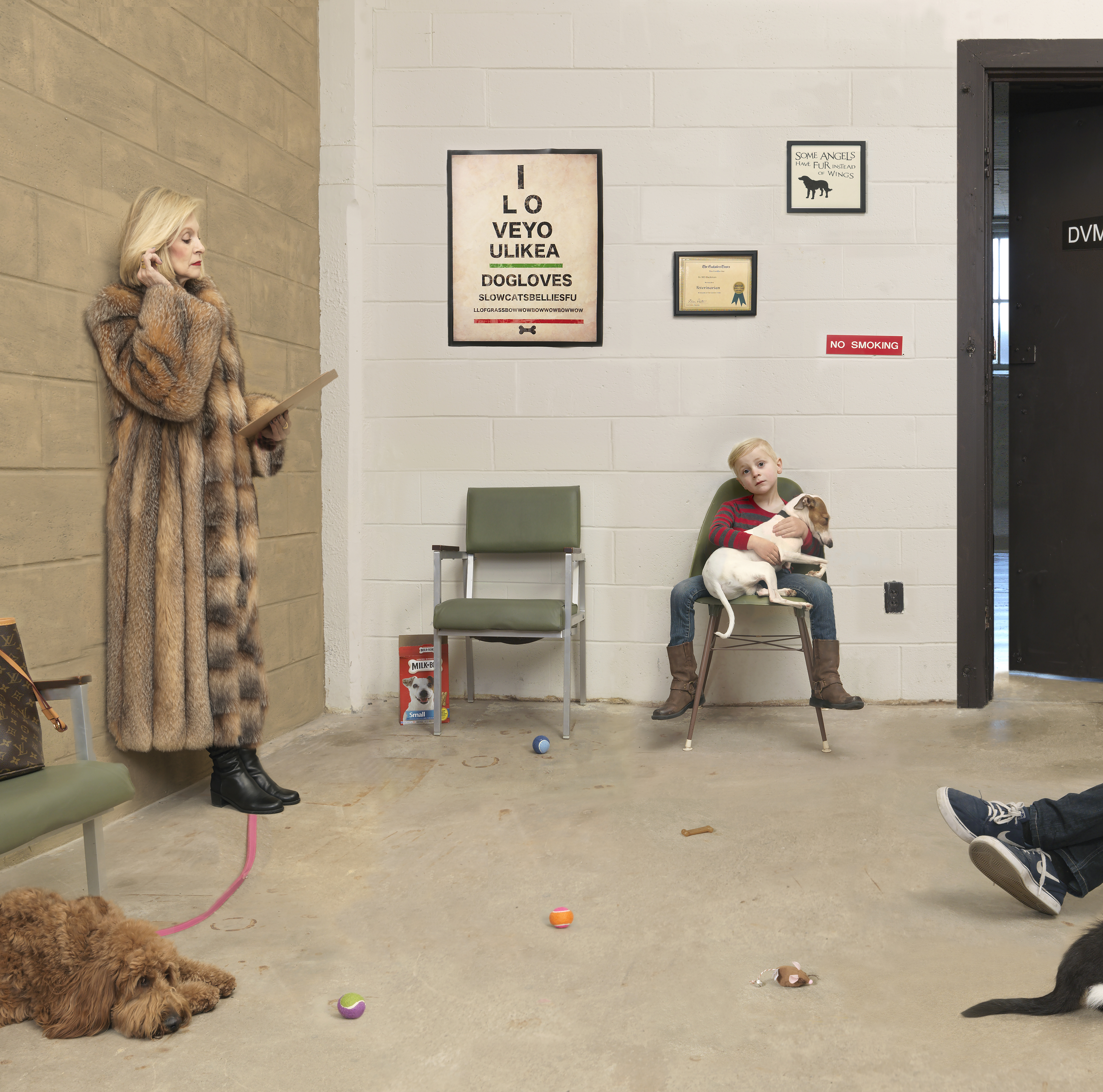A woman in a fur coat stands beside a man in dark shoes with legs stretched out on the floor, while a young boy sits holding a small dog in a green chair. Dogs and toys scatter across a concrete floor in a clinical waiting room with white brick walls, framed dog-themed posters, and a "No Smoking" sign.