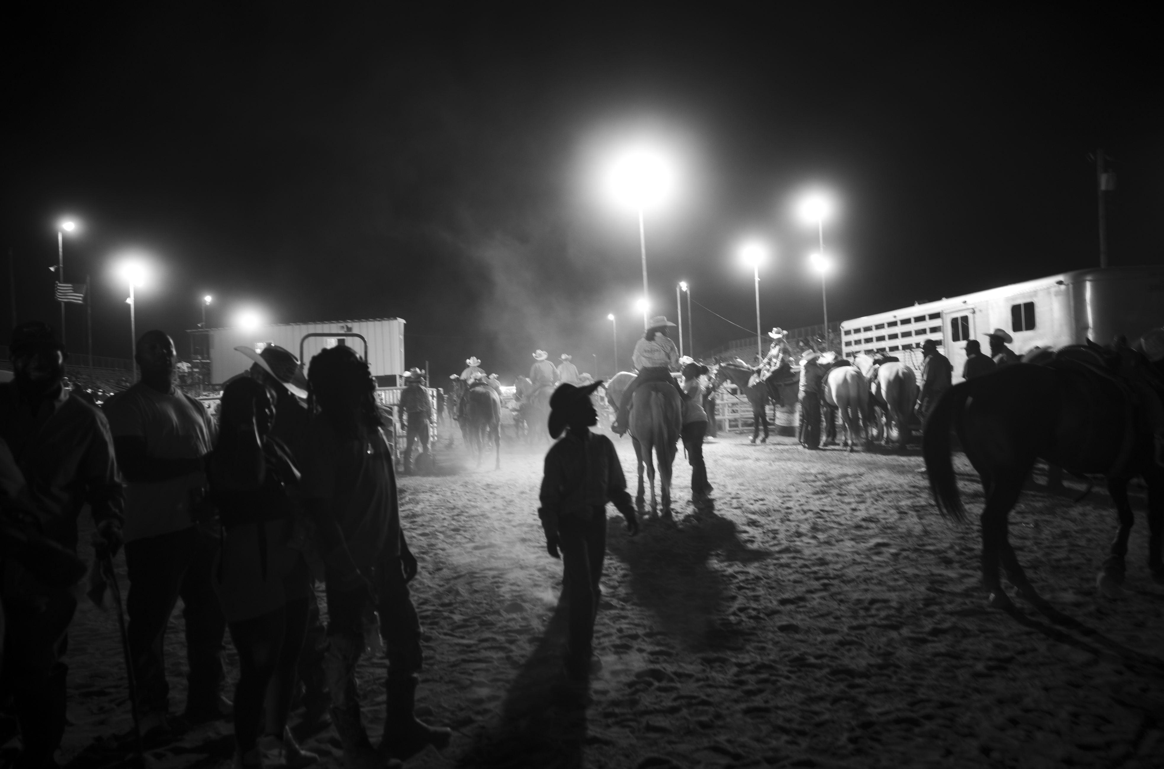 A black and white photograph captures a nighttime gathering in a sandy arena or rodeo grounds, illuminated by bright overhead lights. Numerous figures in silhouette stand scattered across the ground, with horses and what appears to be temporary structures visible in the background, creating an ethereal, atmospheric nocturnal scene.