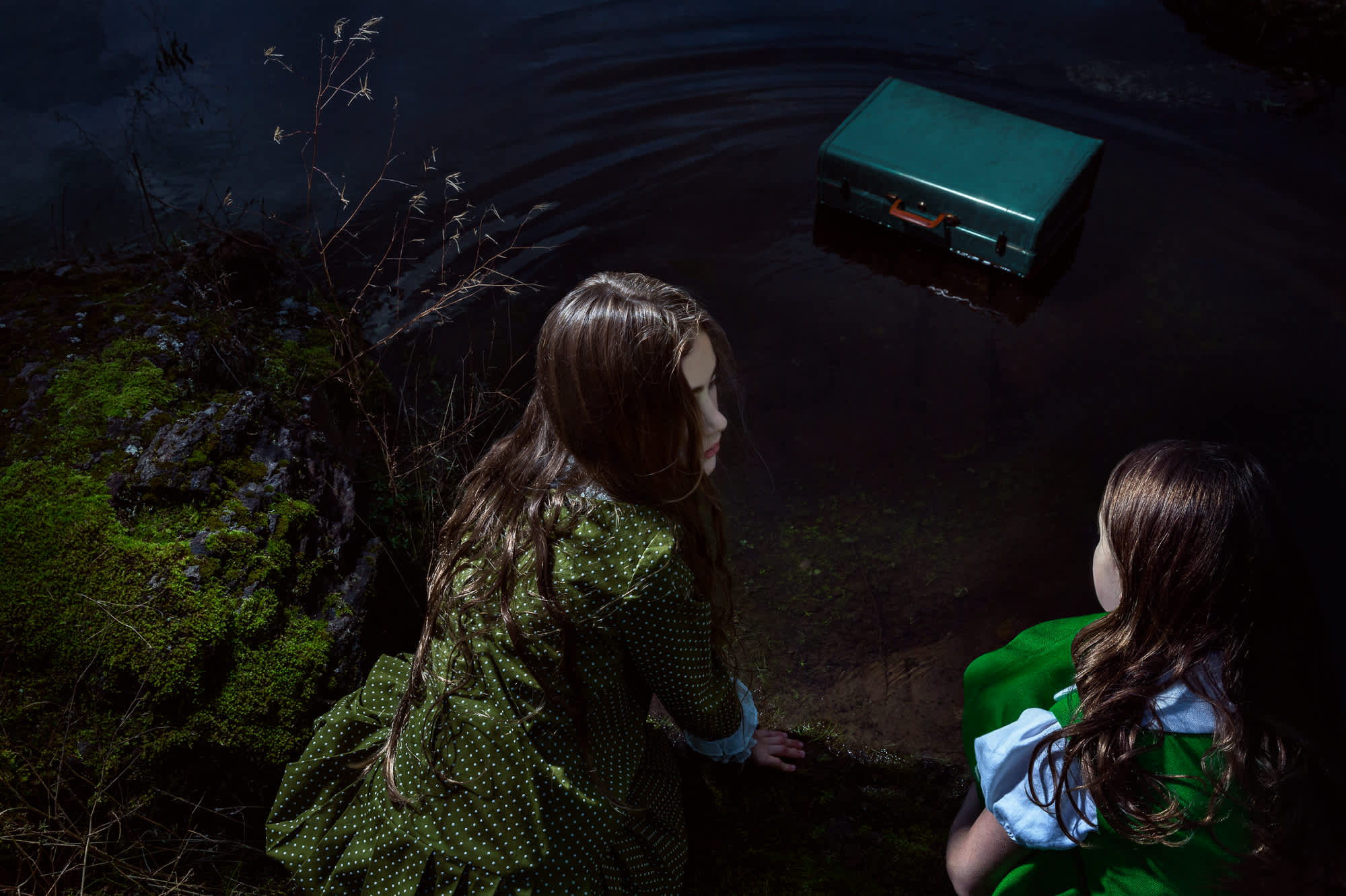 Two young girls in green jackets stand at the edge of dark water surrounded by moss-covered rocks and sparse vegetation, gazing toward a turquoise-colored box floating on the water's surface.