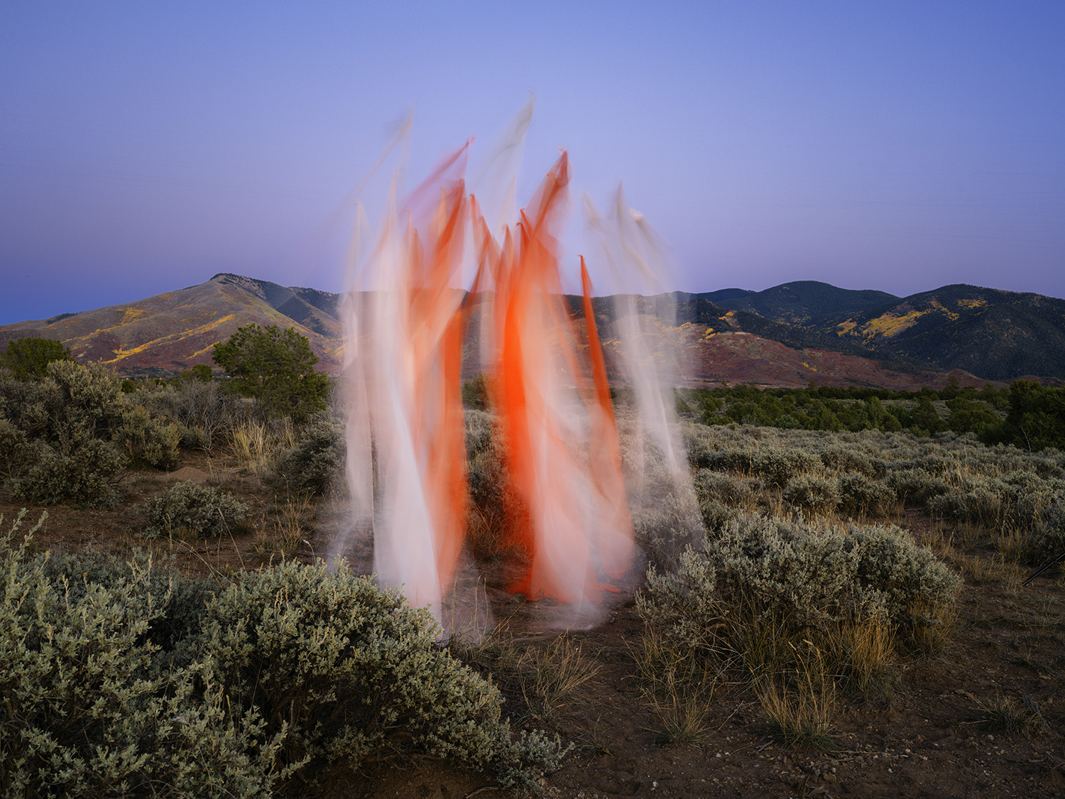 Billowing white and orange silk fabric suspended in a desert landscape, creating dramatic vertical forms against a twilight sky. Arid hills dotted with sparse vegetation frame the ephemeral installation.