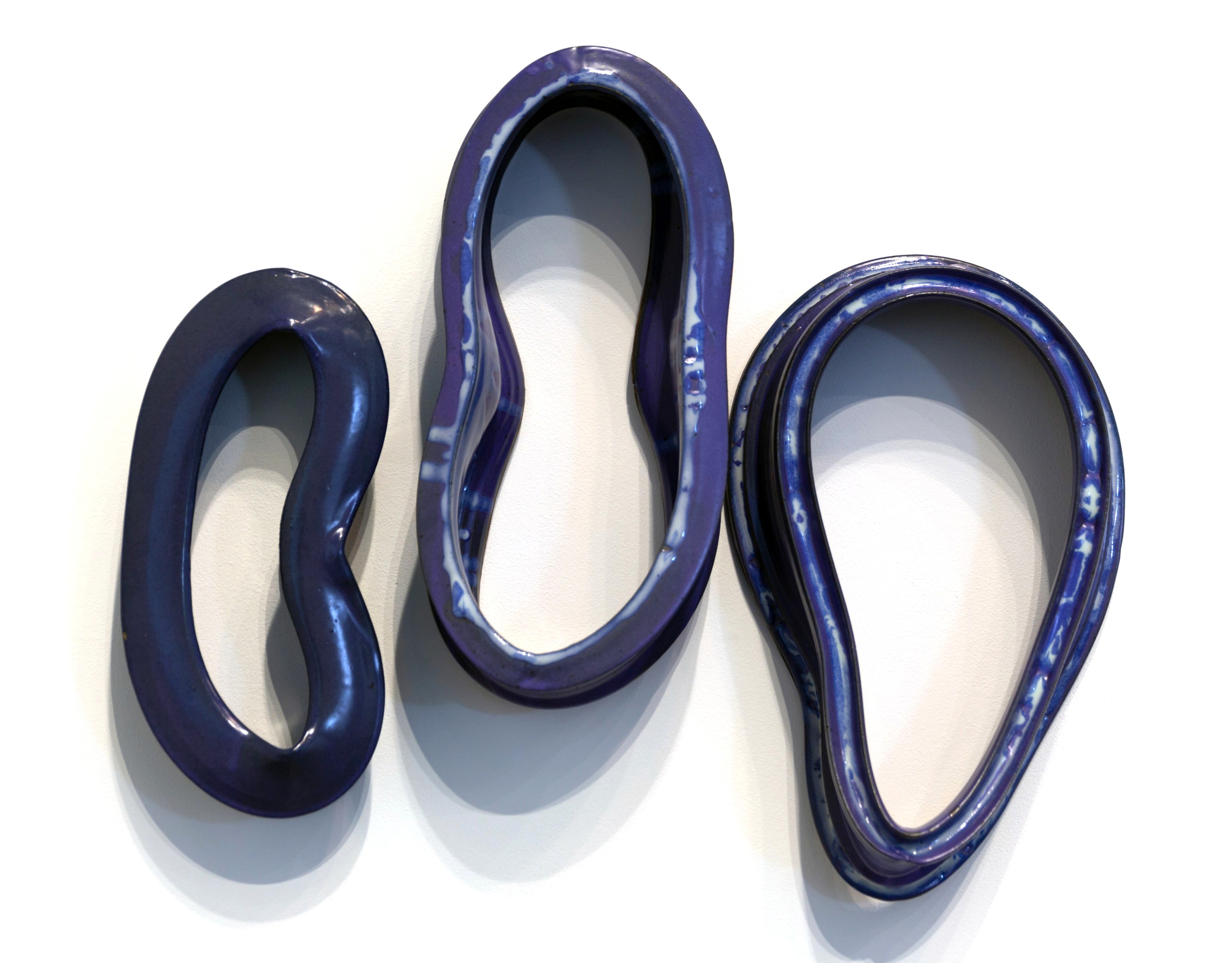 Three deep blue ceramic rings of varying sizes arranged in a row against a white wall, each casting a soft shadow. The glossy, glazed surfaces reflect light, revealing the organic, hand-formed quality of the sculptural objects.
