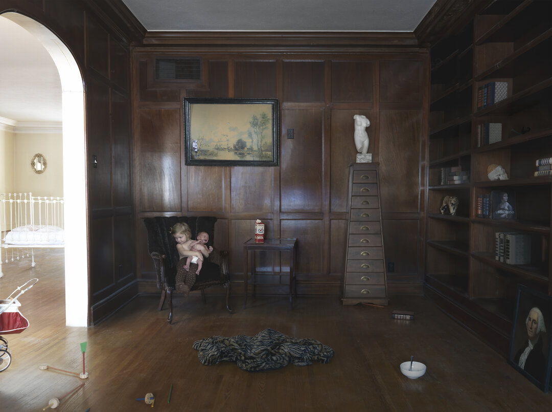 A richly paneled wood-lined interior room features a woman seated in a black chair holding a small child, surrounded by scattered toys and domestic objects on a polished wooden floor. Shelving displays sculptures, books, and portraits, while natural light streams through an arched doorway, creating a staged domestic scene that blends everyday life with gallery-like presentation.