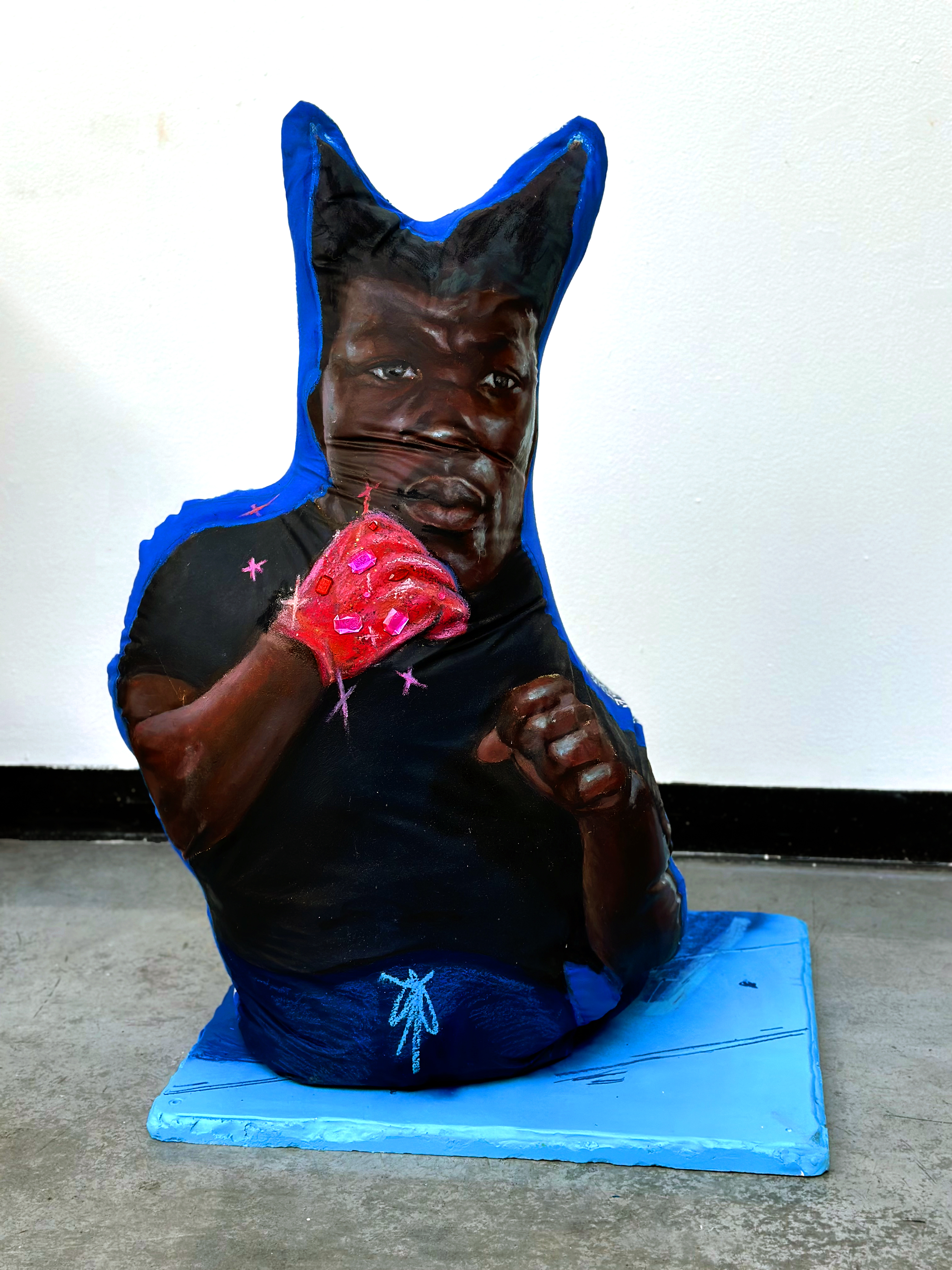 A sculptural figure with an exaggerated cat-like head in deep brown sits on a bright blue rectangular base. The figure wears a black shirt and blue pants, with one arm raised displaying a vivid red, textured glove adorned with pink embellishments. Small pink stars are scattered across the black garment, while a delicate blue figure sketch appears on the pants leg.