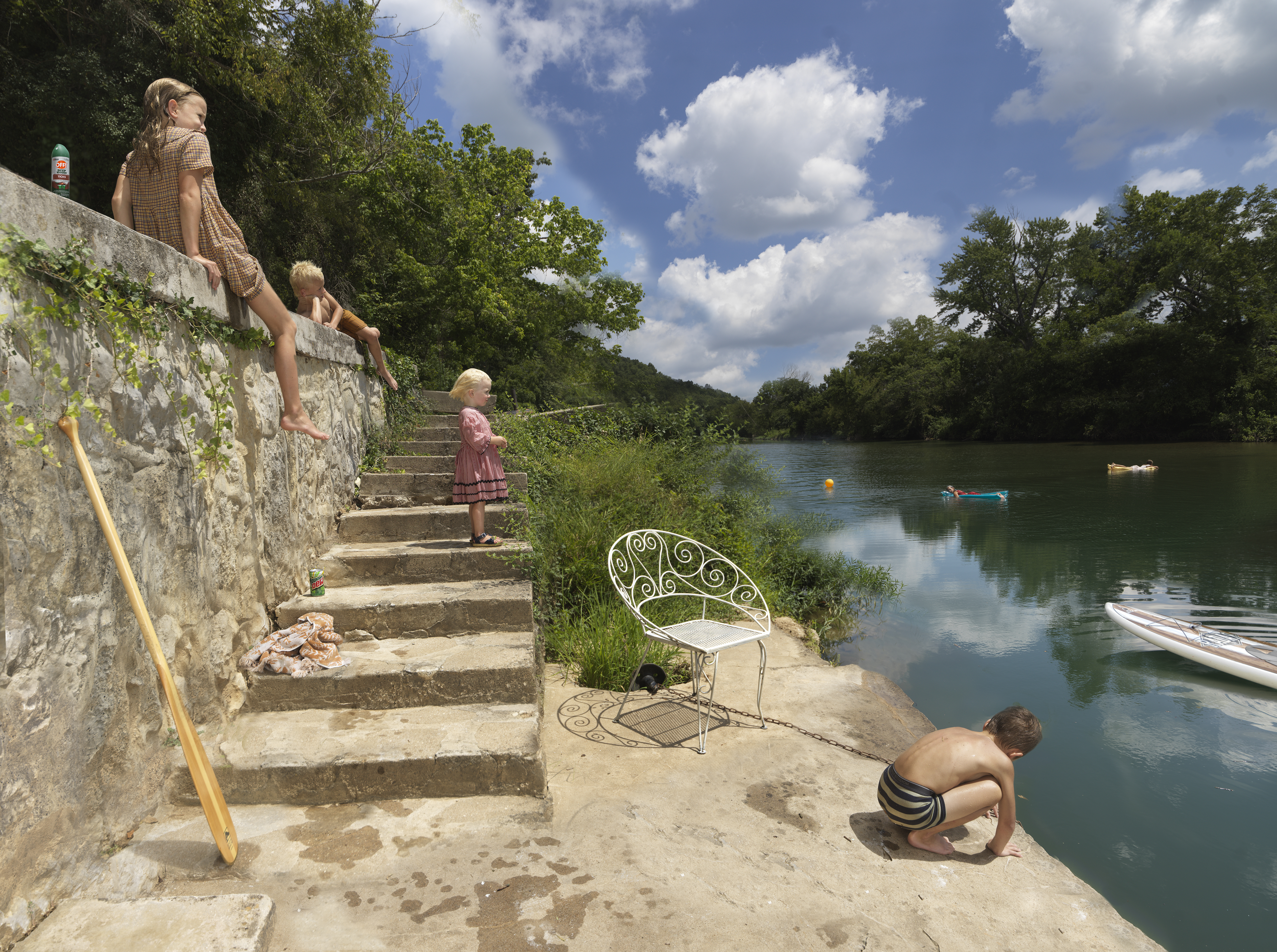 Children play on stone steps descending to a river, where others swim and paddle boats. A decorative wrought-iron chair sits empty on the sandy beach, while lush green trees frame the peaceful waterside scene under a partly cloudy sky.