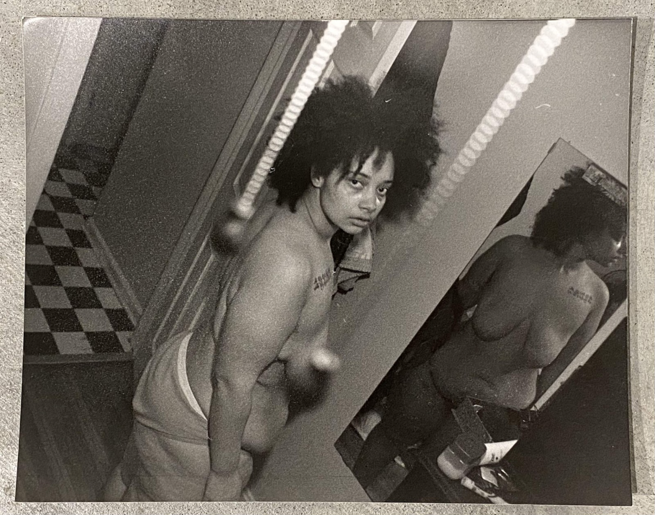 Black and white photograph showing two shirtless figures in an interior space with fluorescent ceiling lights and patterned flooring. The figure on the left, with voluminous dark hair, gazes toward the camera with a contemplative expression, while a second figure is visible on the right side of the frame. The composition captures an intimate domestic moment rendered in stark, high-contrast tones characteristic of silver gelatin printing.