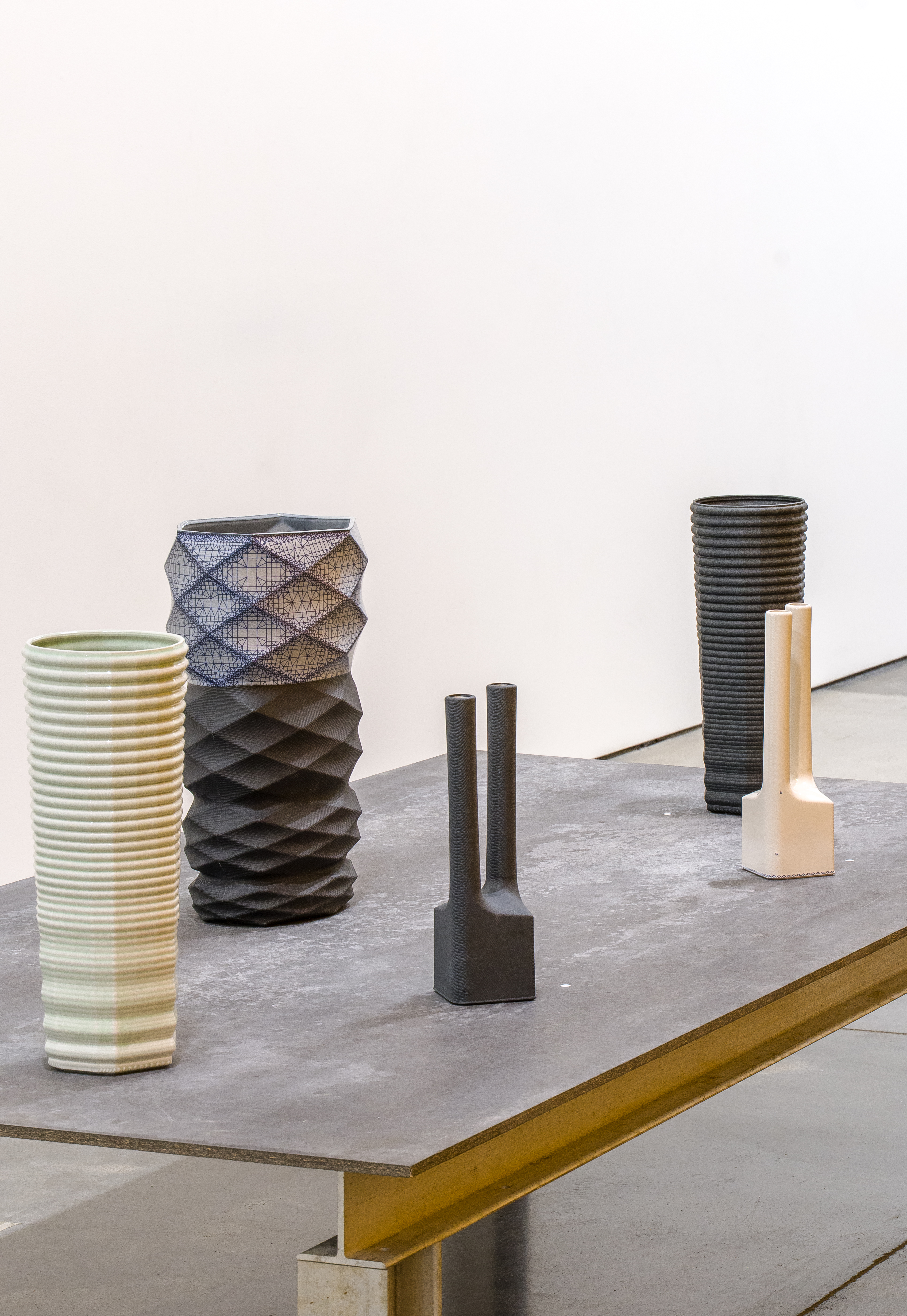 Five sculptural ceramic objects arranged on a concrete-topped table with a brass edge. From left to right: a ribbed cylindrical vessel in cream and sage green; a textured dark vessel with blue mesh inlay and quilted surface pattern; two small dark cylindrical forms; and a tall ridged black cylinder paired with a cream-colored geometric base. The compositions demonstrate varied surface treatments, textures, and forms arranged in a gallery setting against a neutral background.