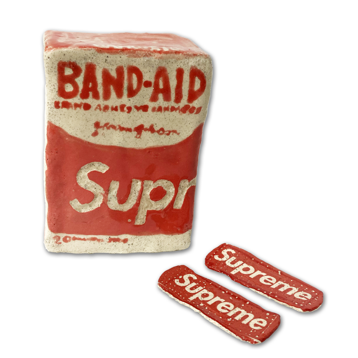 A ceramic sculpture of a vintage Band-Aid box rendered in cream and red glazed clay, accompanied by two small matching ceramic Band-Aid strips. The box features period typography and branding details in relief, with a weathered, aged appearance suggesting both the original product design and the materiality of handcrafted clay.