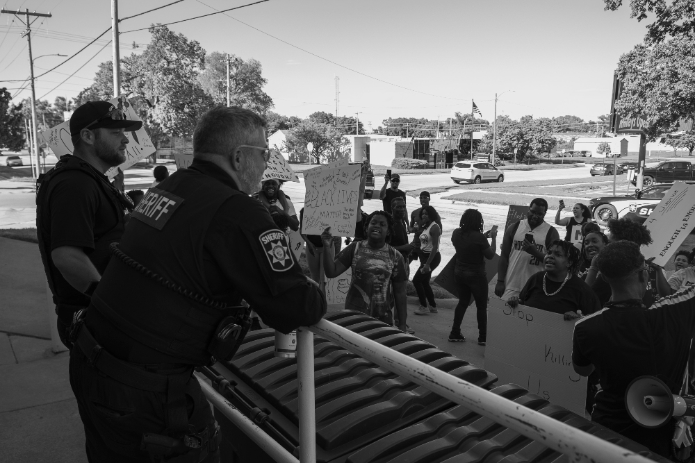 Black and white photograph of a street protest scene showing law enforcement officers in uniform standing near a metal railing, facing a crowd of demonstrators holding signs. Trees, power lines, and commercial buildings are visible in the background under an overcast sky.