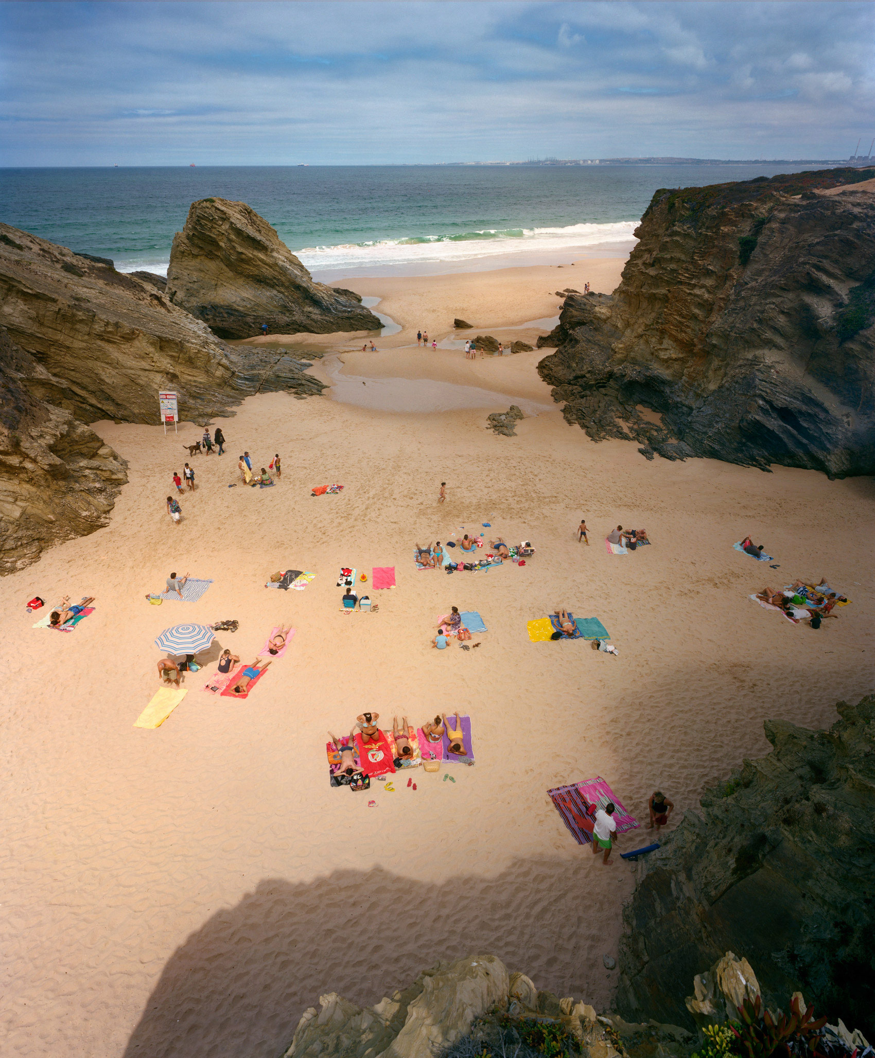 Aerial view of a sandy beach nestled between dramatic rocky outcrops, with scattered beachgoers, colorful umbrellas, and towels dotting the shore. Turquoise waters and white-capped waves frame the cove, while a clouded sky stretches across the horizon.