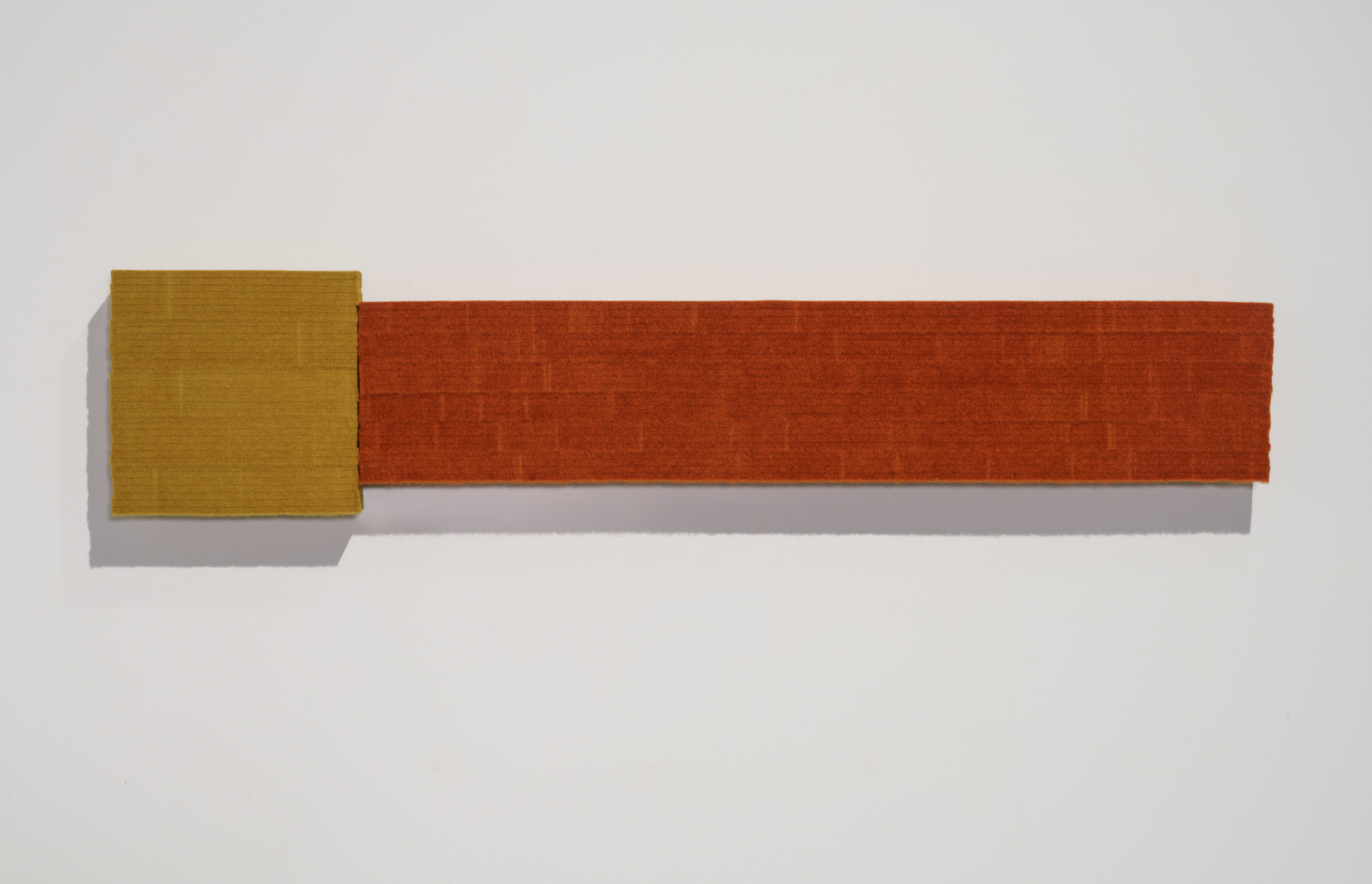 Two rectangular panels of industrial felt joined horizontally—a smaller ochre-yellow square on the left and a longer rust-orange rectangle extending to the right—mounted on a white wall, casting soft shadows beneath.