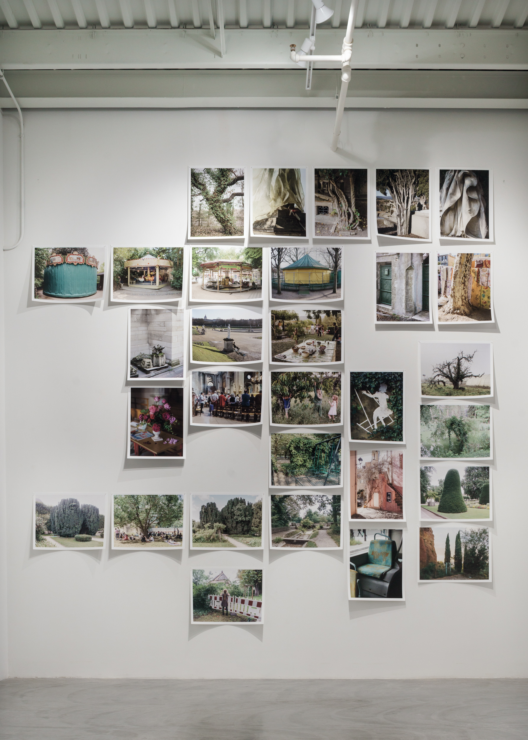 A gallery wall installation of approximately forty color photographs arranged in an organic, scattered pattern against white walls. The prints, mounted with white borders in the style of instant film, depict French parks, gardens, architecture, trees, and public spaces, interspersed with images of carousels, garden furniture, and intimate domestic scenes. The composition creates a rhythmic, constellation-like arrangement across the expansive wall surface.