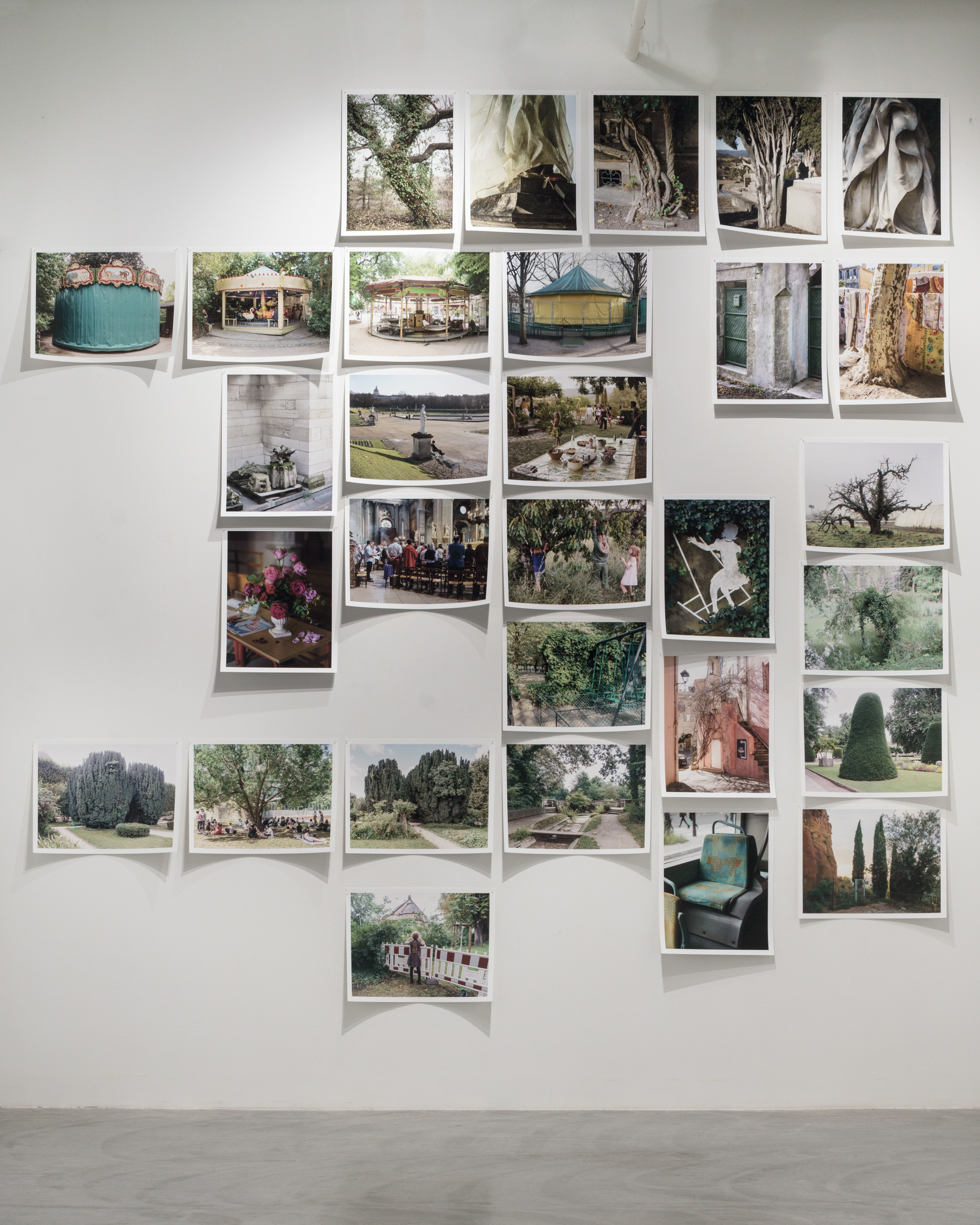 A white gallery wall displays approximately forty Polaroid-style photographs arranged in an organic, scattered composition. The photographs document French landscapes, architectural details, gardens, and public spaces, featuring recurring motifs of trees, historic structures, and verdant grounds rendered in natural colors of greens, earth tones, and weathered materials. The prints cast subtle shadows against the wall, creating depth and dimension across the installation.