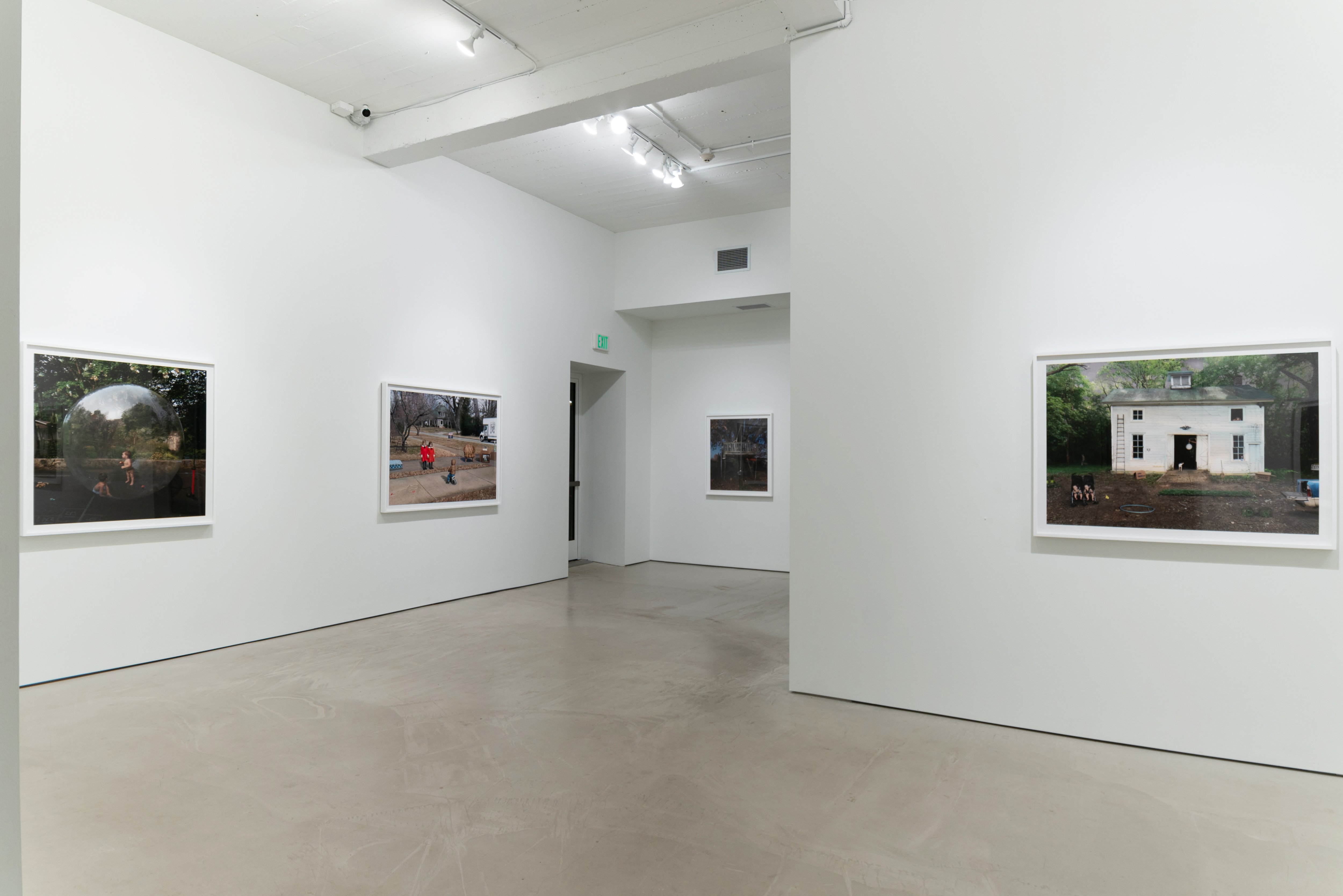 A bright, minimalist gallery interior with polished concrete flooring displays four large framed color photographs hung on white walls. The images depict domestic and outdoor scenes featuring people in naturalistic settings. Track lighting illuminates the space, with an open doorway visible in the center background.
