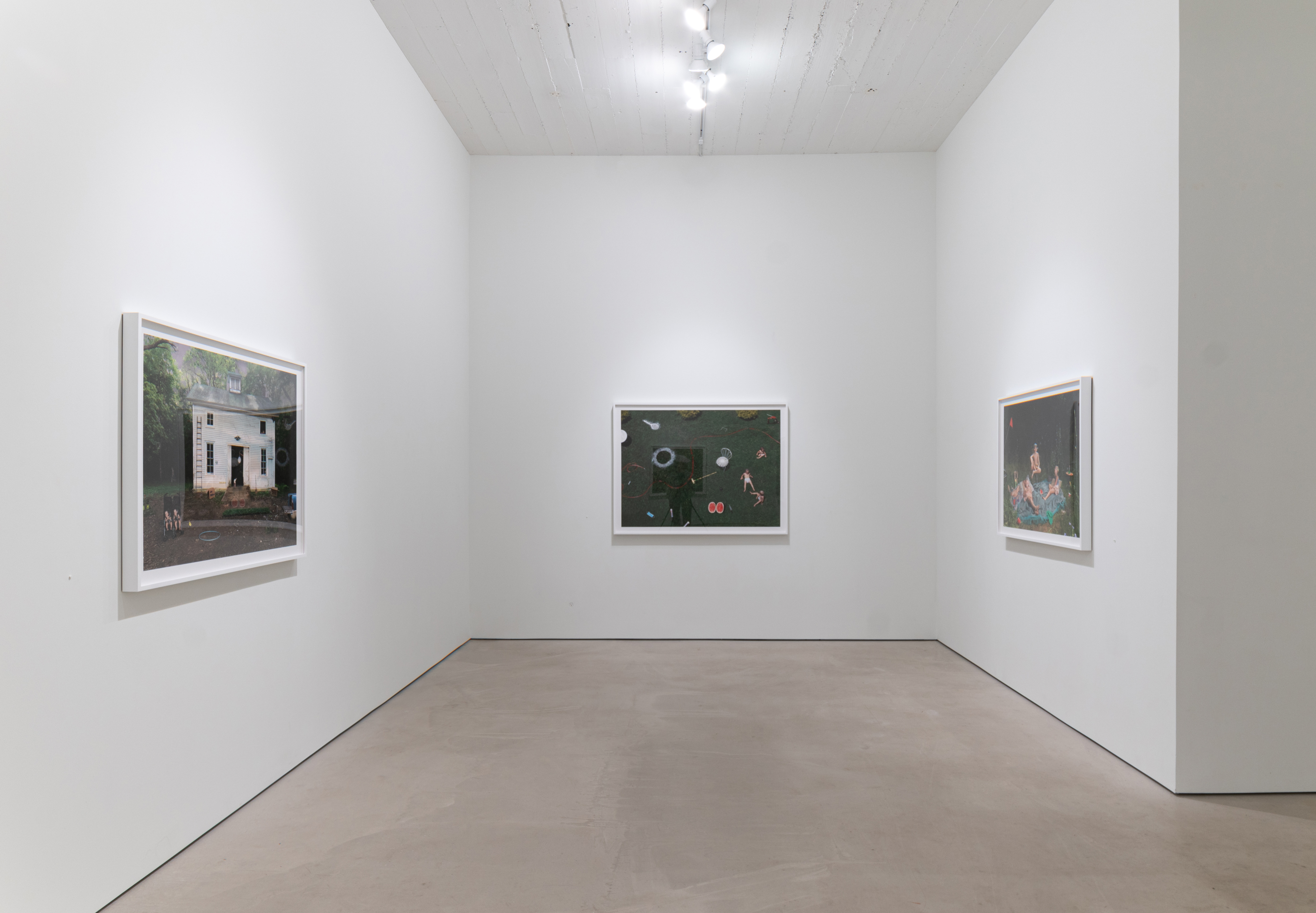 Three framed color photographs hung on white gallery walls: at left, a small white house with figures in front; center, a dark forest scene with floating objects and figures; at right, a richly colored nocturnal gathering of figures. Polished concrete floor, track lighting overhead.