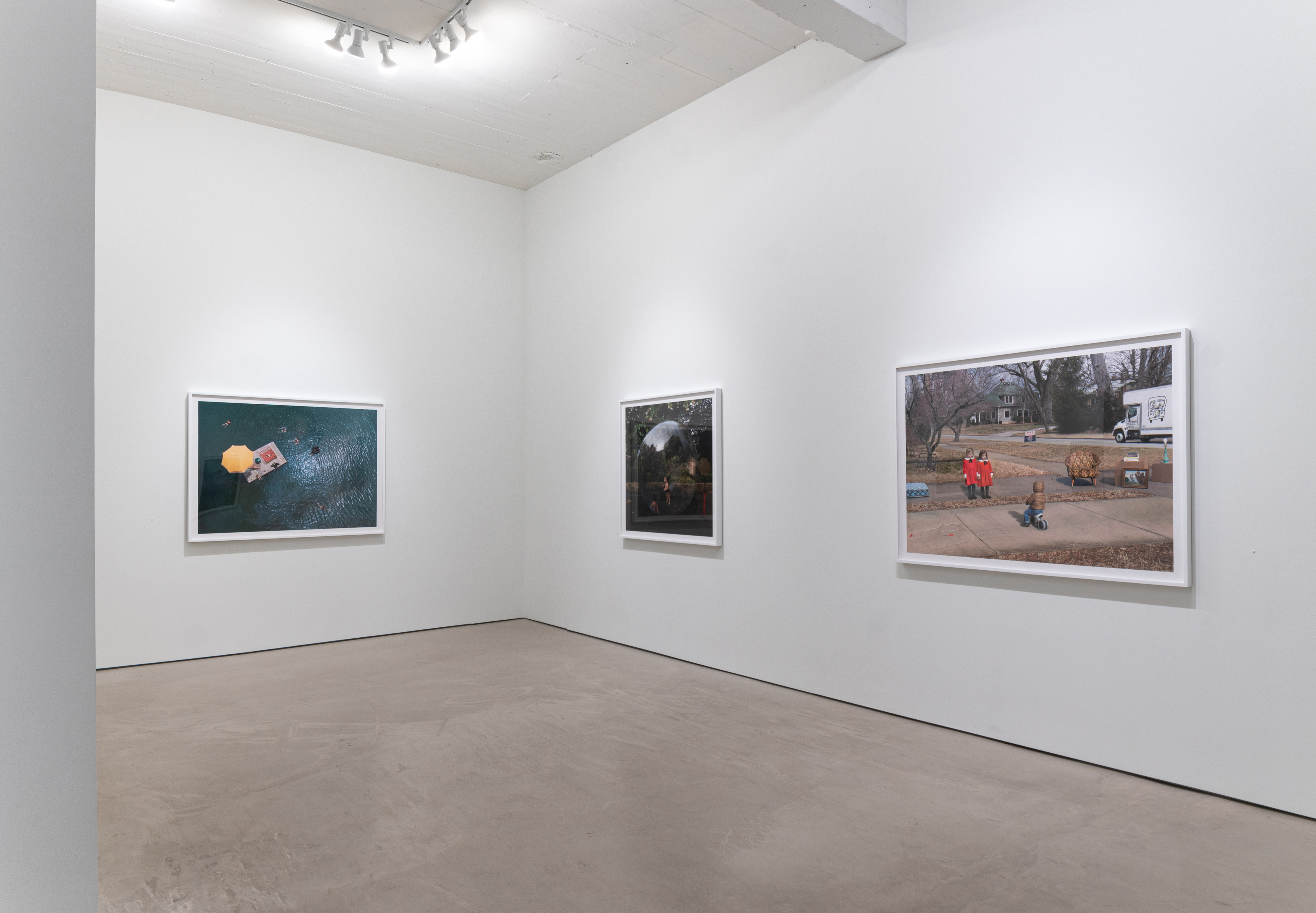 Three framed color photographs of varying sizes hang on white gallery walls in a minimalist installation. The left image depicts an aerial view of a person in a yellow inner tube on dark water; the center shows a nighttime scene with figures and atmospheric lighting; the right displays a daytime suburban scene with children playing in a dirt yard near a house and parked vehicle.