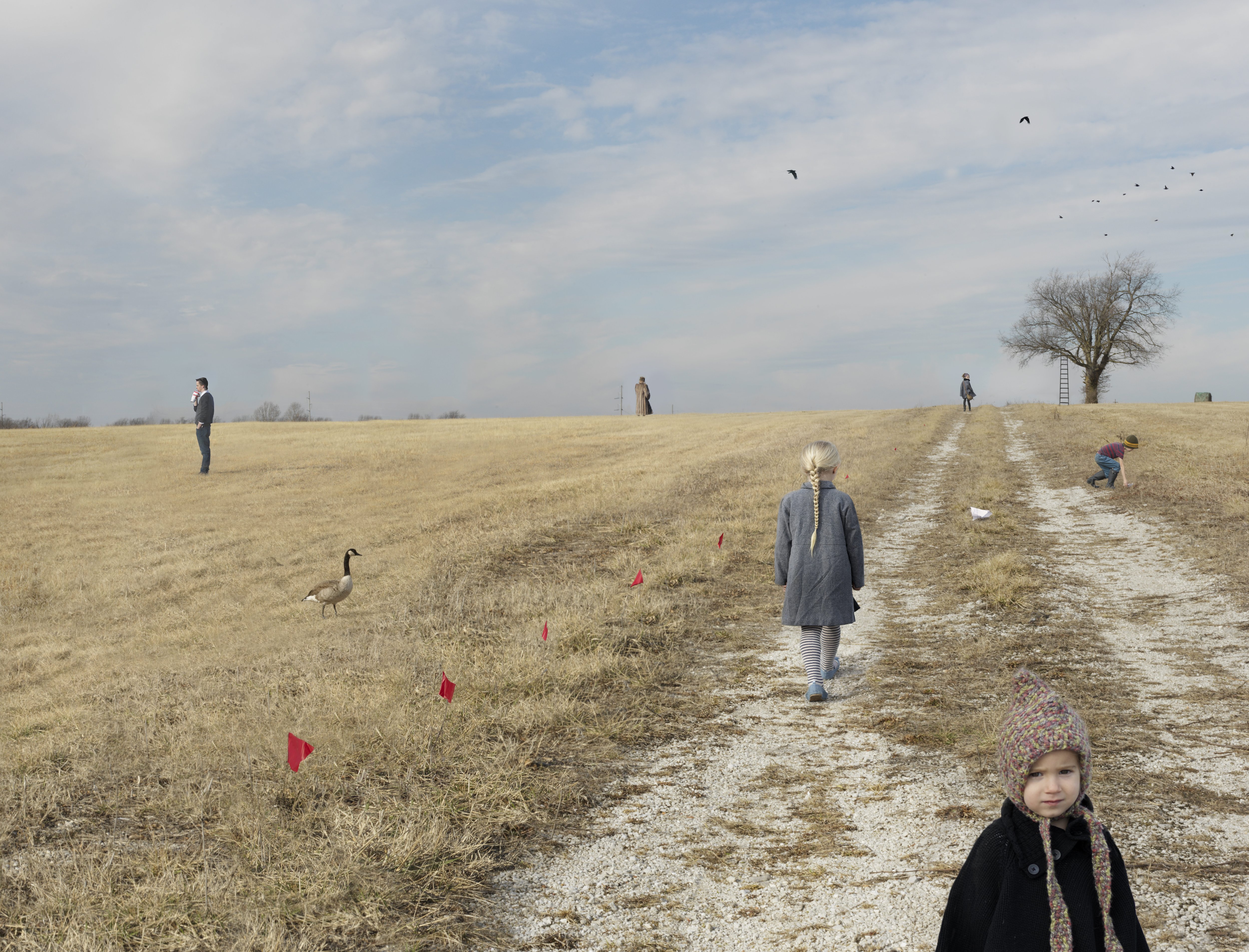 A sprawling rural landscape with sparse dry grass and a dirt path stretching toward the horizon. Multiple figures of varying ages are scattered across the field, some walking along the path and others standing in the grassland, with red flags marking the ground. A bare tree stands in the distance, and a goose wanders in the foreground. The overcast sky and muted earth tones dominate the composition, creating an ambiguous, contemplative scene.