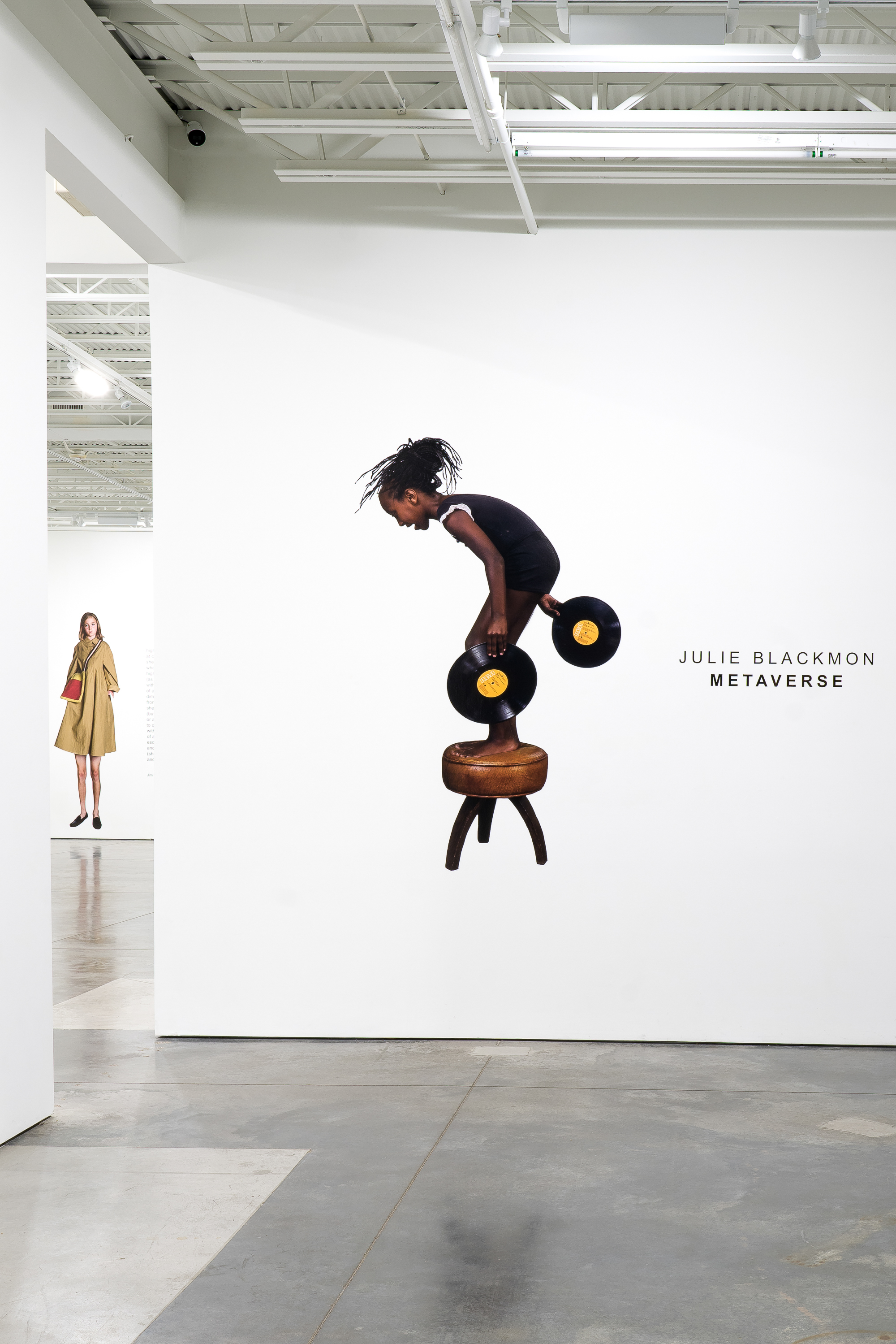 A figure in a black sleeveless top performs an acrobatic pose atop a wooden stool with three legs, balancing while holding black vinyl records with gold centers in each hand. The minimalist gallery setting features white walls, polished concrete flooring, and industrial ceiling with exposed beams. On the left wall, a fashion photograph shows a woman wearing a mustard-yellow coat.