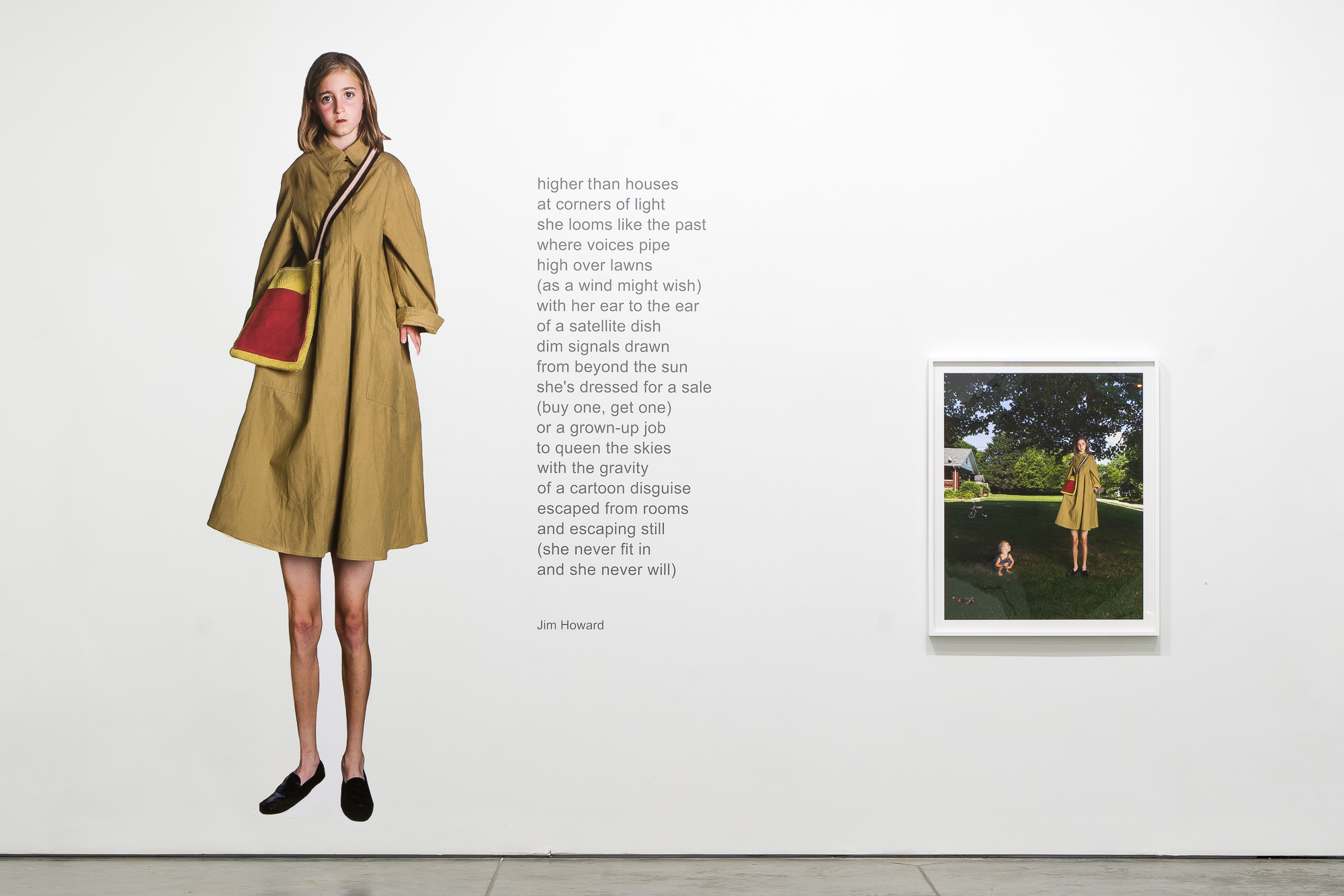 Gallery wall installation featuring a life-size photograph of a young woman in a mustard-colored coat with a red bag standing against a white wall, paired with an framed photograph showing the same figure in a yellow dress standing in a grassy yard. Gray text displaying a poem by Jim Howard is positioned between the two images.