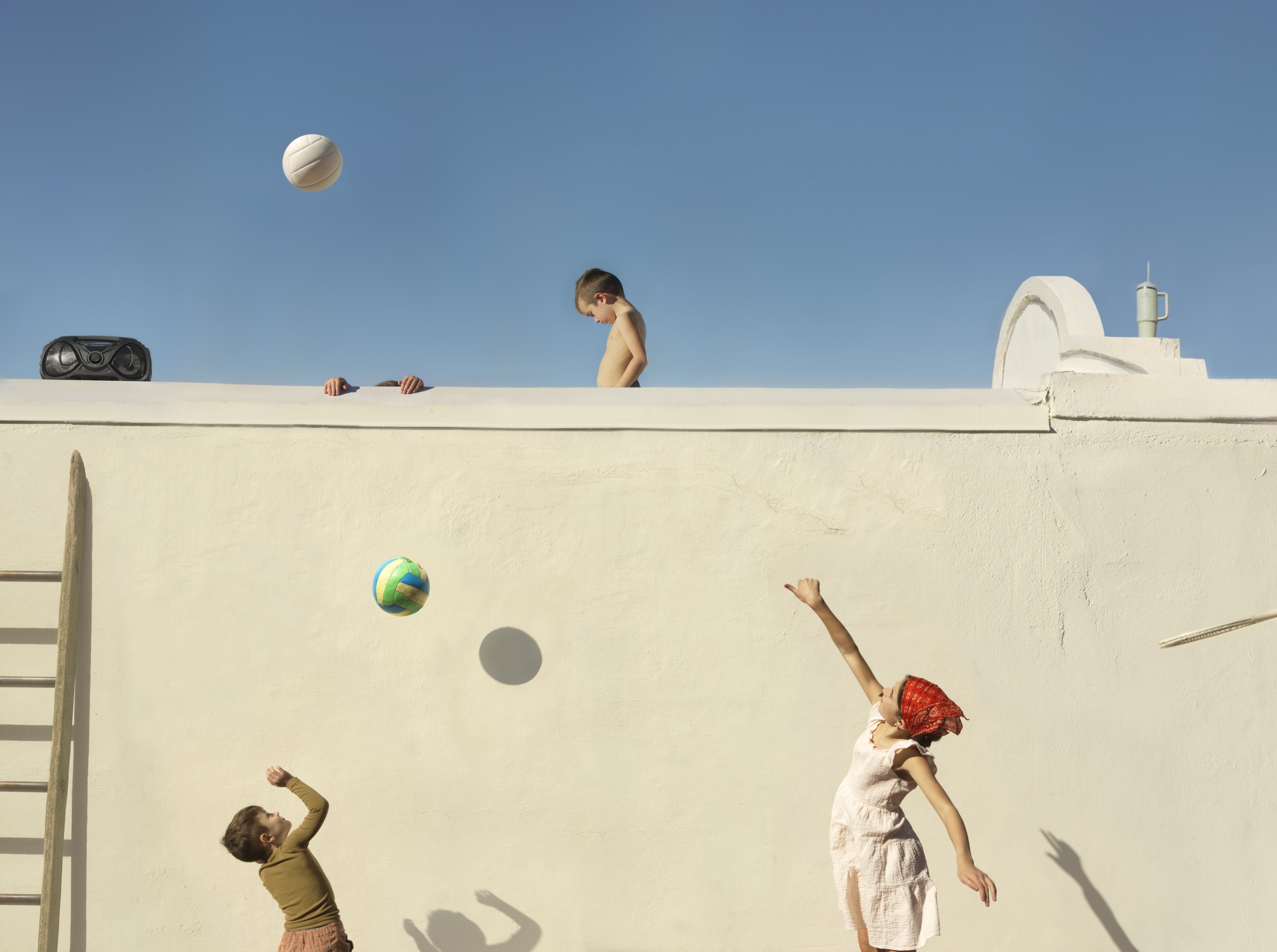 Children play with balls against a white concrete wall, with one child perched on top and others positioned below. The composition is divided between the bright blue sky above and the stark white architectural surface, creating a striking contrast that emphasizes the playful figures and their shadows cast on the wall.