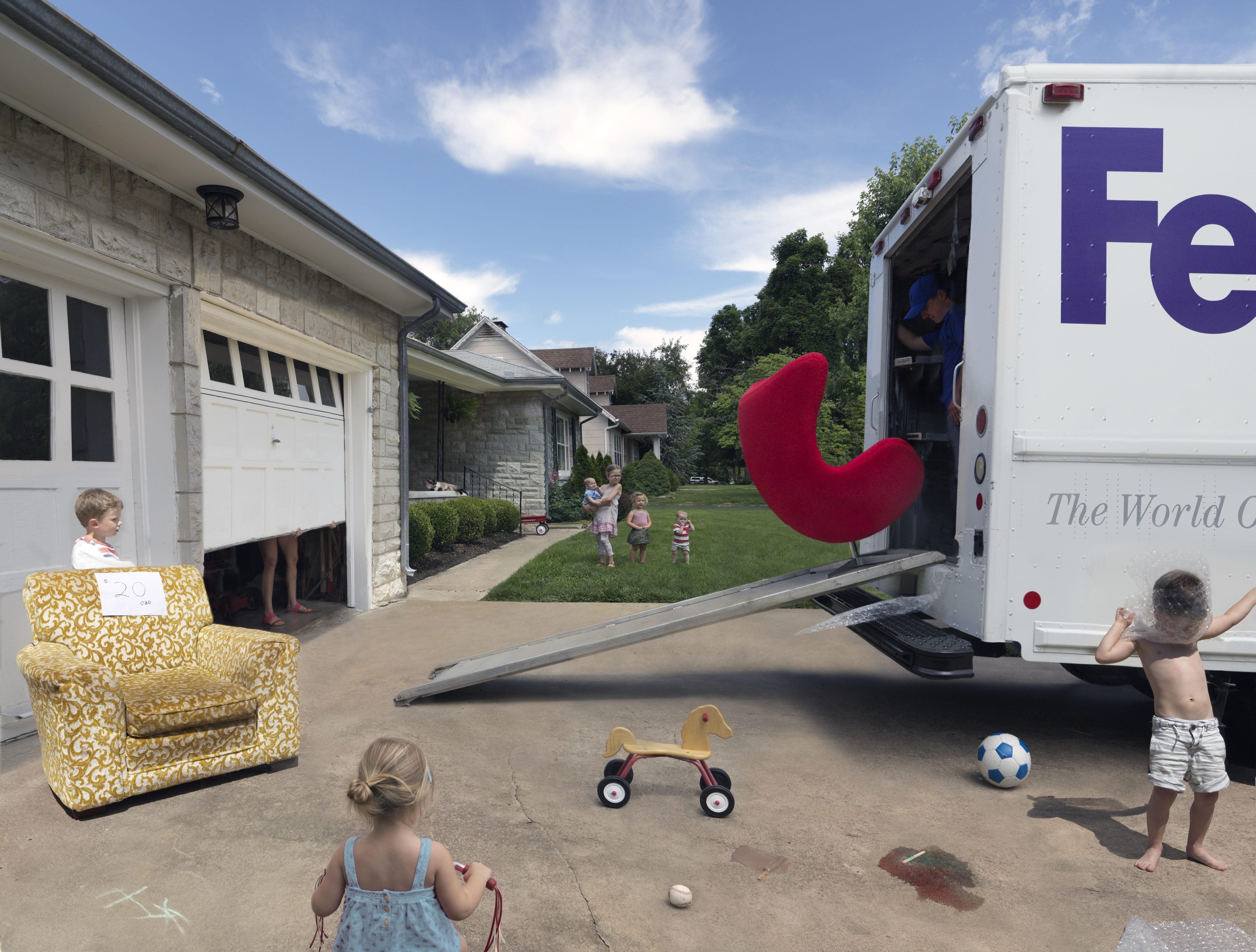 A suburban driveway scene with children playing among oversized objects: a giant yellow patterned armchair on the left, a large red curved form mounted on a delivery truck on the right, and scattered toys including a wooden duck-shaped rider and soccer ball. Residential houses and green lawn visible in the background under a blue sky.