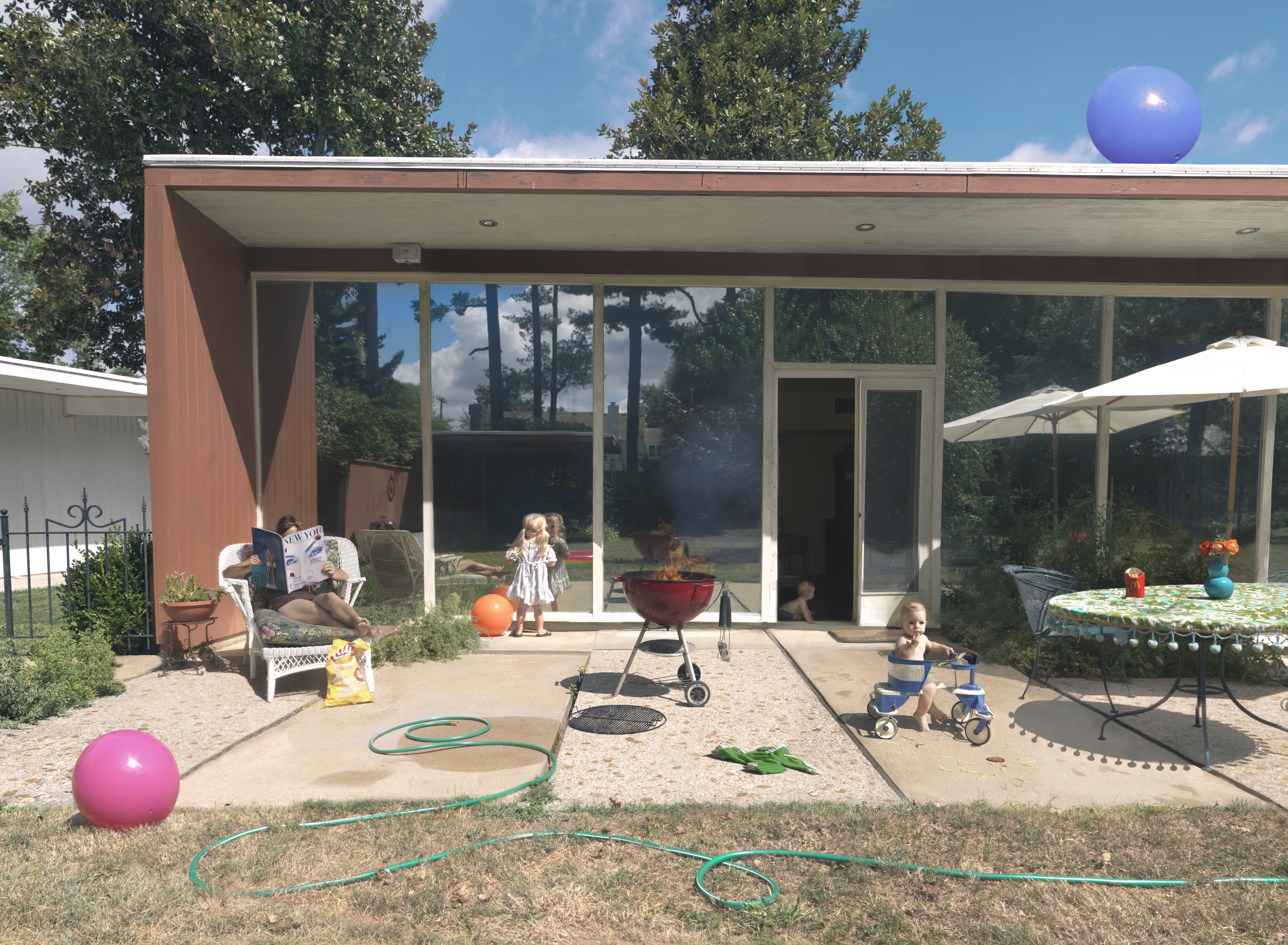 A contemporary residential patio scene depicts multiple figures of varying ages engaged in leisure activities. The composition features a modernist house with expansive glass doors and rust-colored metal cladding, a red kettle grill, scattered toys including colorful balls and a tricycle, garden hoses, umbrellas, and patio furniture arranged across a sandy ground. The bright daylight and clear sky create strong shadows and vibrant color contrasts throughout the domestic landscape.