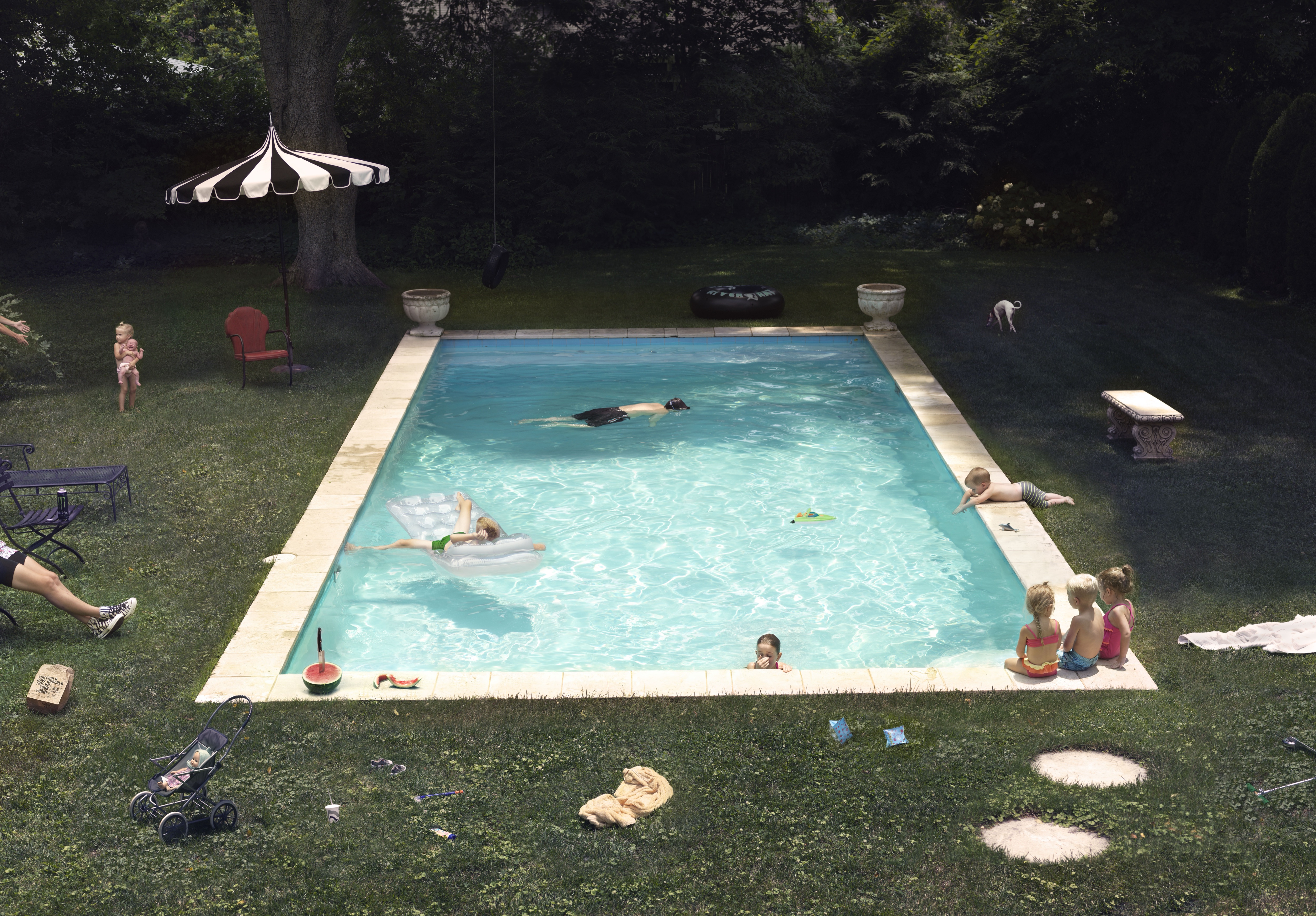 A rectangular swimming pool filled with turquoise water dominates a manicured lawn, surrounded by children and adults engaged in various leisure activities. A striped black and white umbrella shelters a red chair on the left, while figures float, swim, and sit poolside, with decorative urns, garden furniture, and a natural tree canopy creating an idealized suburban summer scene.
