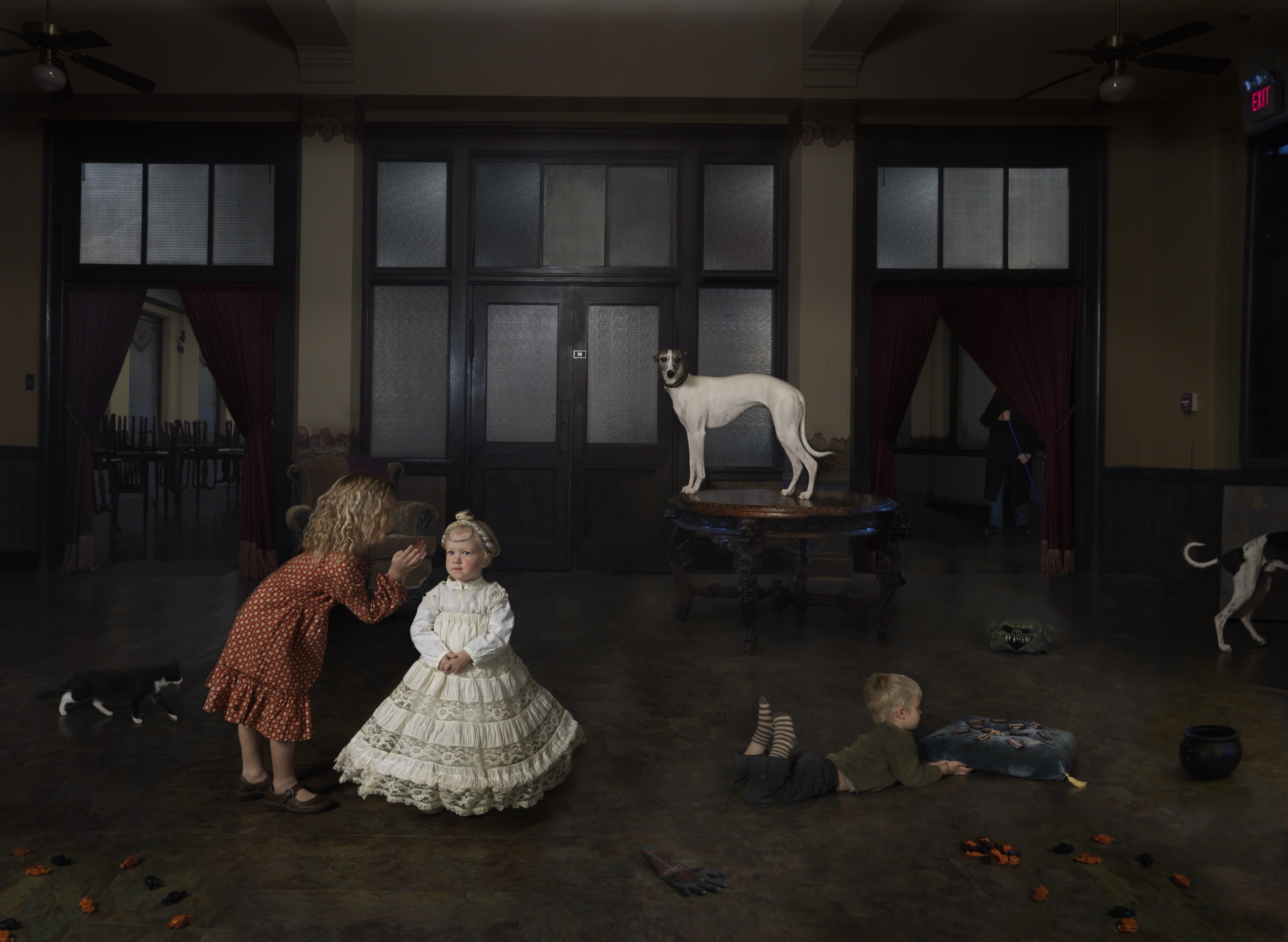A meticulously composed interior scene depicts children in period dress interacting with multiple dogs in a modernist gallery space with dark-framed glass doors and frosted panels. A child in an ornate white dress stands center while another in a rust-colored coat bends toward her; additional children play on the polished floor as white and spotted dogs occupy various positions throughout the room, creating a surreal, theatrical tableau.