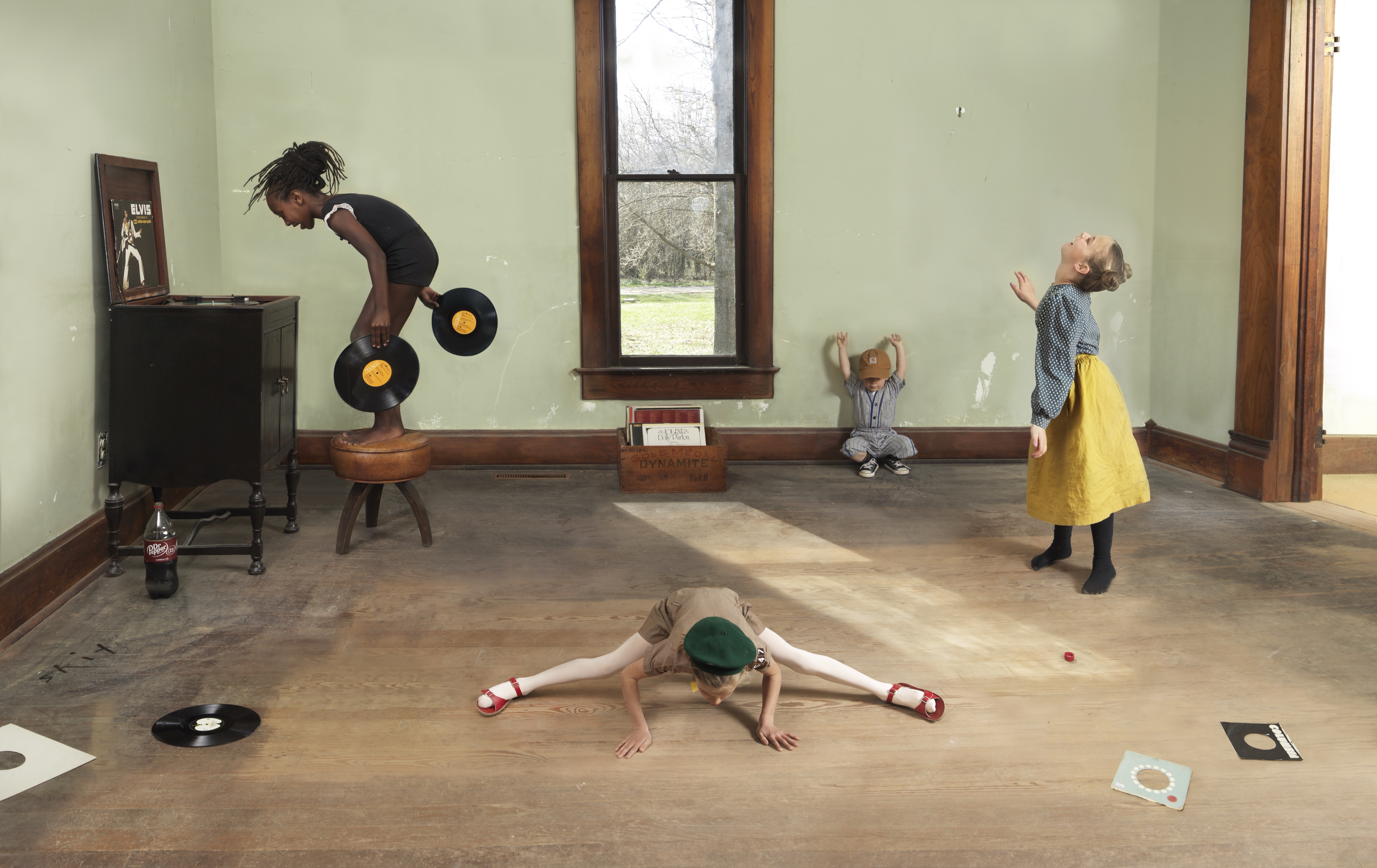 Four figures interact with vinyl records in a sparse gallery space with pale green walls and wooden trim. A man bends forward holding two records above his shoulders, a child in a yellow skirt reaches upward, another child sits on the floor in a split position wearing a green hat, and a small figure in the center raises their arms. Vinyl records, a wooden chair, and other objects are scattered across the wooden floor, with natural light streaming through a tall window.
