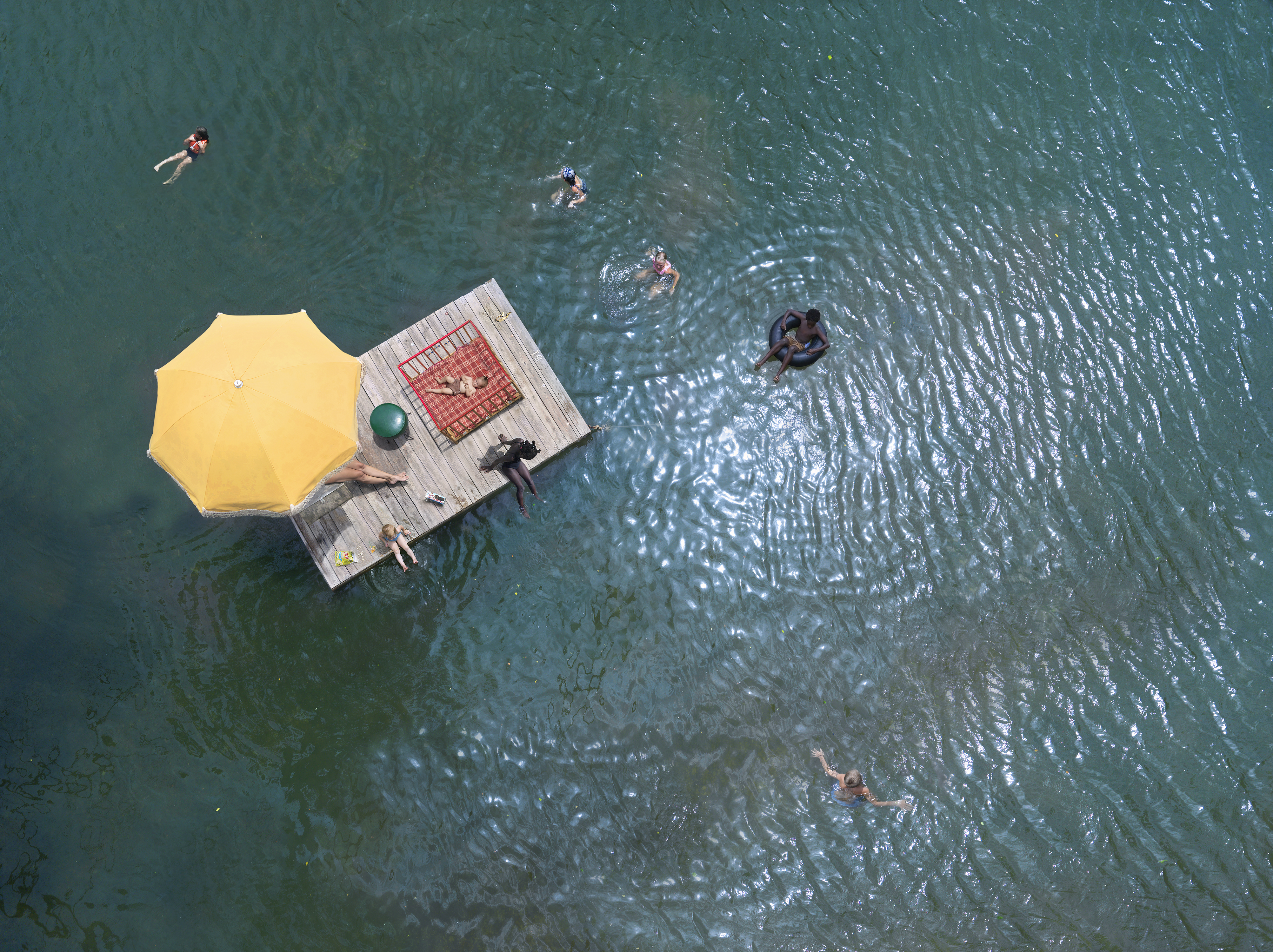 Aerial view of a wooden dock floating on green water, where several people swim and relax. A bright yellow umbrella and a red picnic blanket are set up on the dock's surface, while bathers float and swim in the surrounding water at varying distances from the platform.