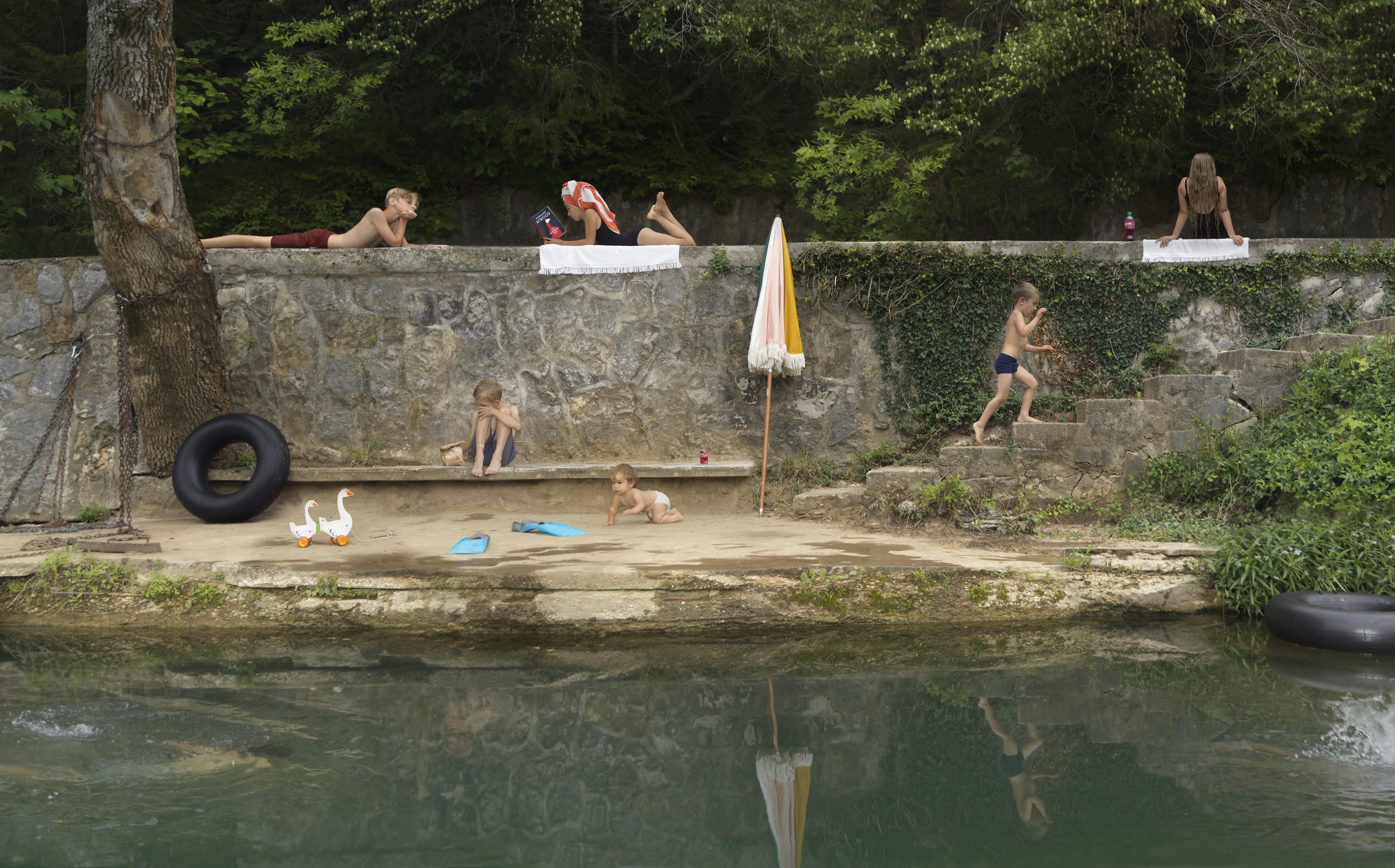 Children and adults relax along a stone riverside embankment on a sunny day, some seated on the wall, others playing in the sandy beach area below. Two geese walk near a black tire and beach items, while an umbrella and towels mark designated spaces, creating a leisurely scene of leisure and recreation framed by dense green foliage above and calm water below.