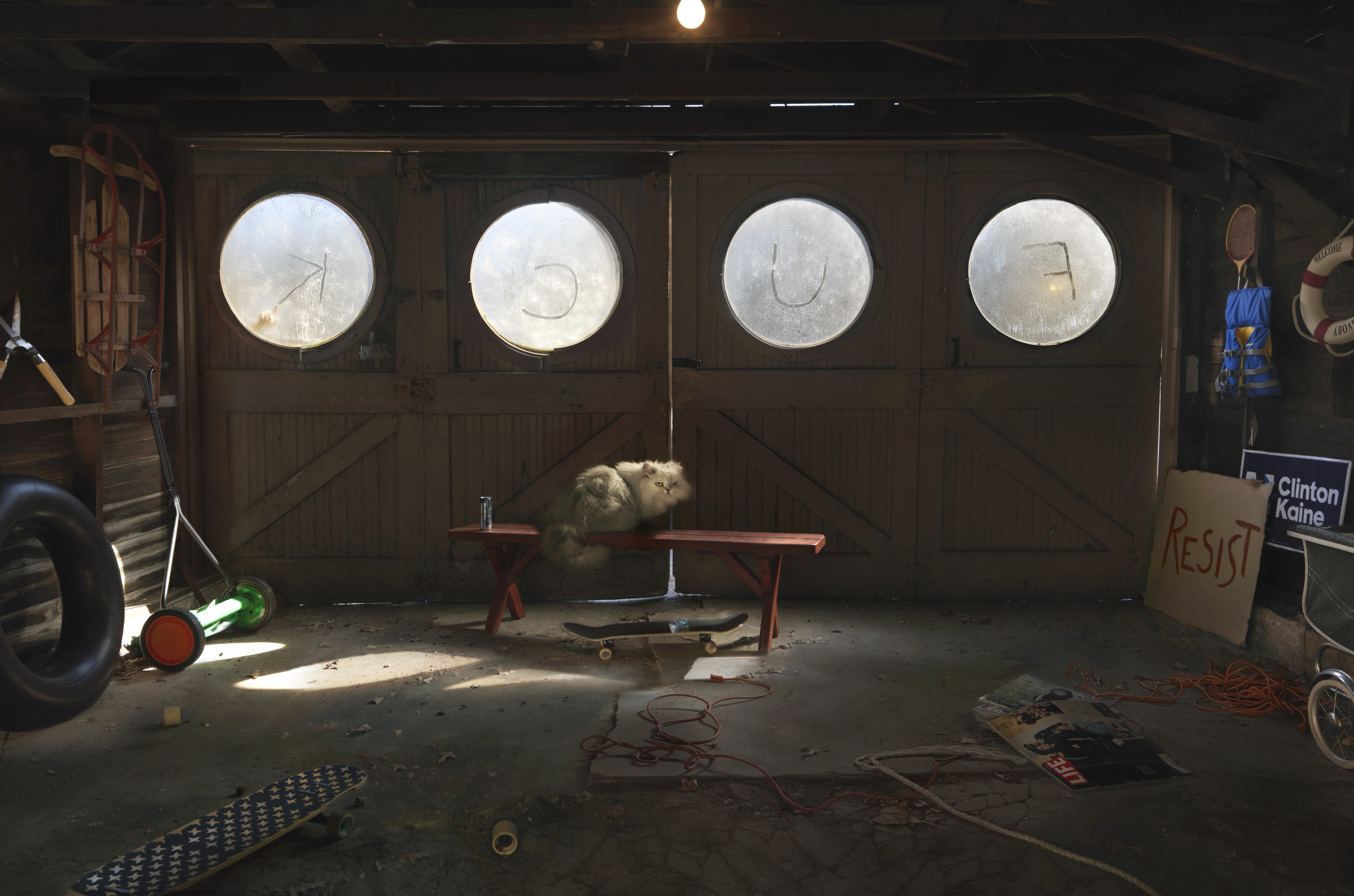 A cluttered garage interior with four large circular windows above wooden doors. Two cats sit on a red bench beneath the windows. Scattered around the concrete floor are various objects including bicycles, a tire, skateboard, tools, electrical cords, and a cardboard sign reading "RESIST" with "Clinton Kaine" visible nearby. Sunlight streams through the circular windows, illuminating the dusty, disorganized space.