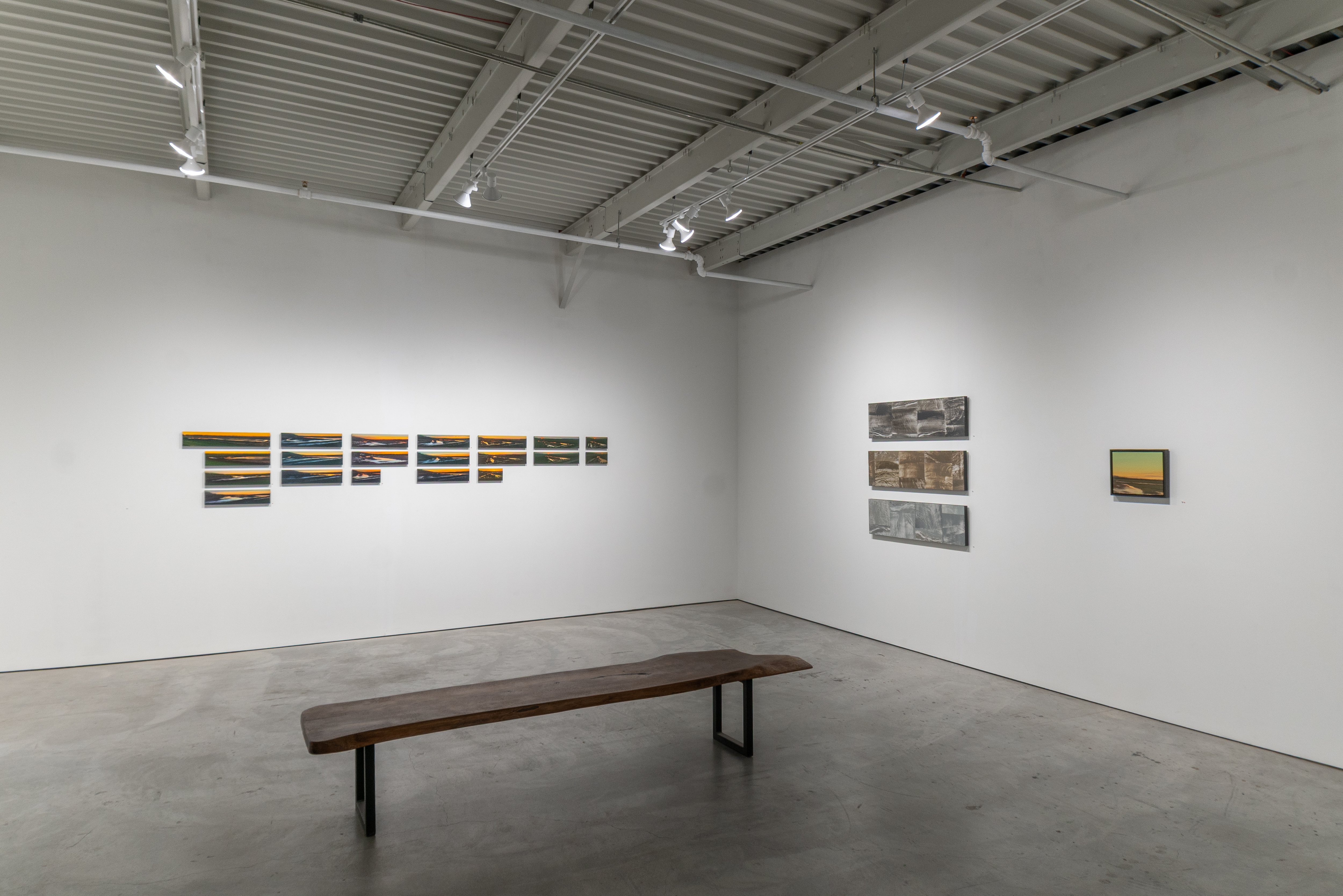 Contemporary gallery interior featuring a wooden bench in the foreground and white walls displaying multiple series of horizontal framed paintings with blue, orange, and gray tones arranged in sequences. Industrial ceiling with exposed metal beams and track lighting.