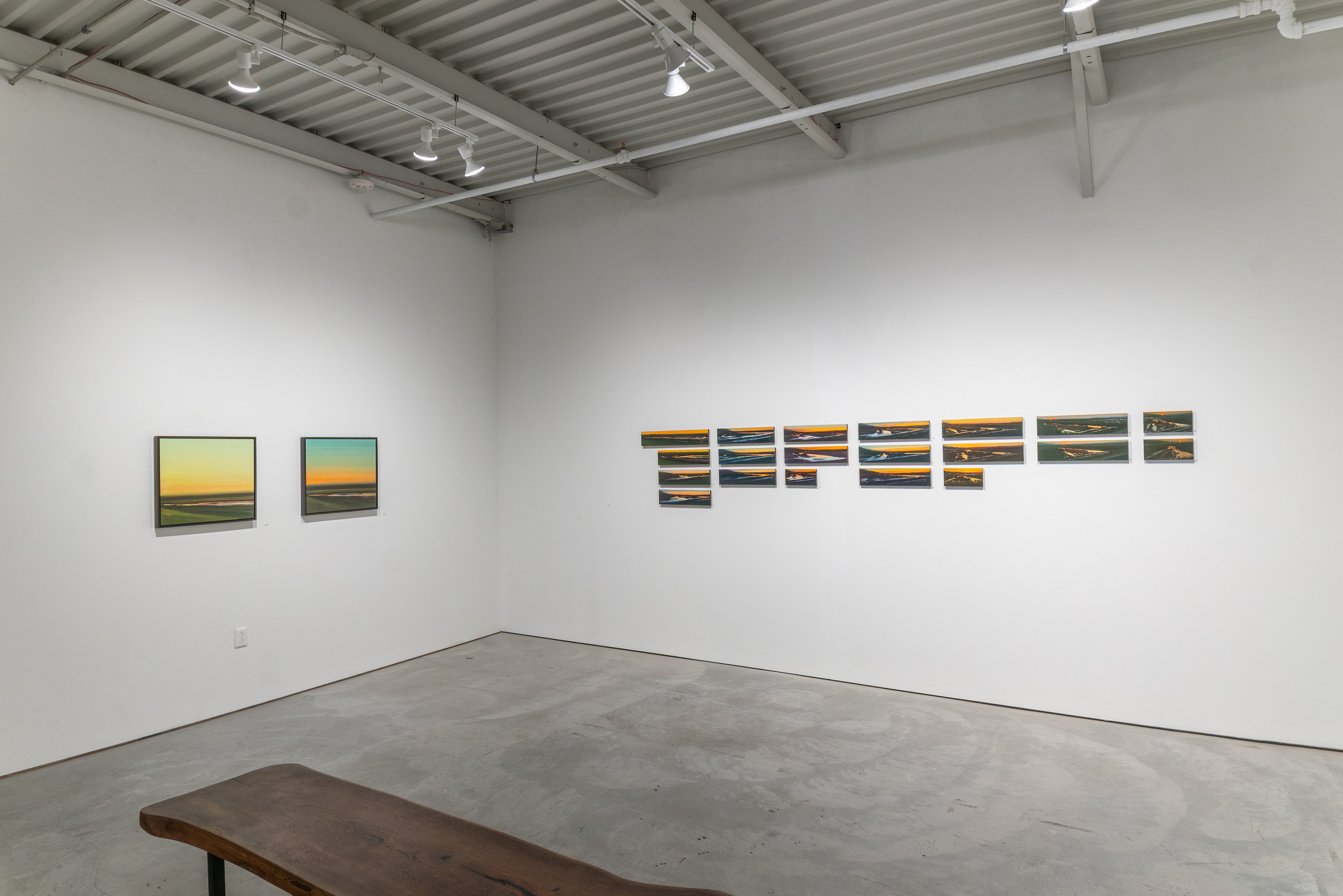 Gallery wall displaying landscape paintings in two distinct groupings: on the left, two framed oil paintings depicting horizons with golden and blue tones; on the right, a series of smaller abstract metallic panels arranged in flowing sequences, featuring gestural marks in gray and bronze tones suggesting water and sand formations. Concrete floor with wooden bench in foreground; industrial ceiling with track lighting.