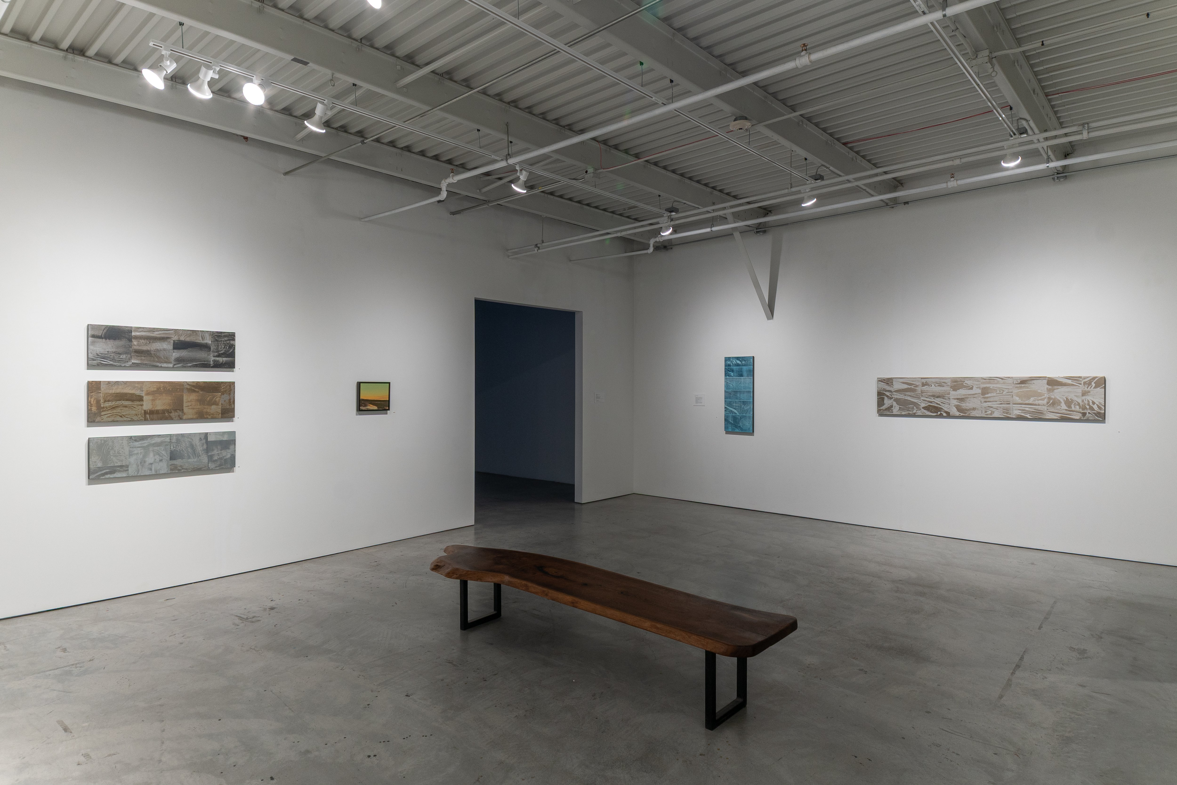 Contemporary art gallery interior with a wooden bench centered on polished concrete flooring. White walls display six paintings of varying sizes arranged asymmetrically—three horizontal abstract compositions on the left in muted earth tones and grays, a small yellow-toned work center-left, and two larger horizontal pieces on the right featuring textured surfaces in beige and blue-gray tones. Industrial ceiling with exposed ductwork and recessed lighting.