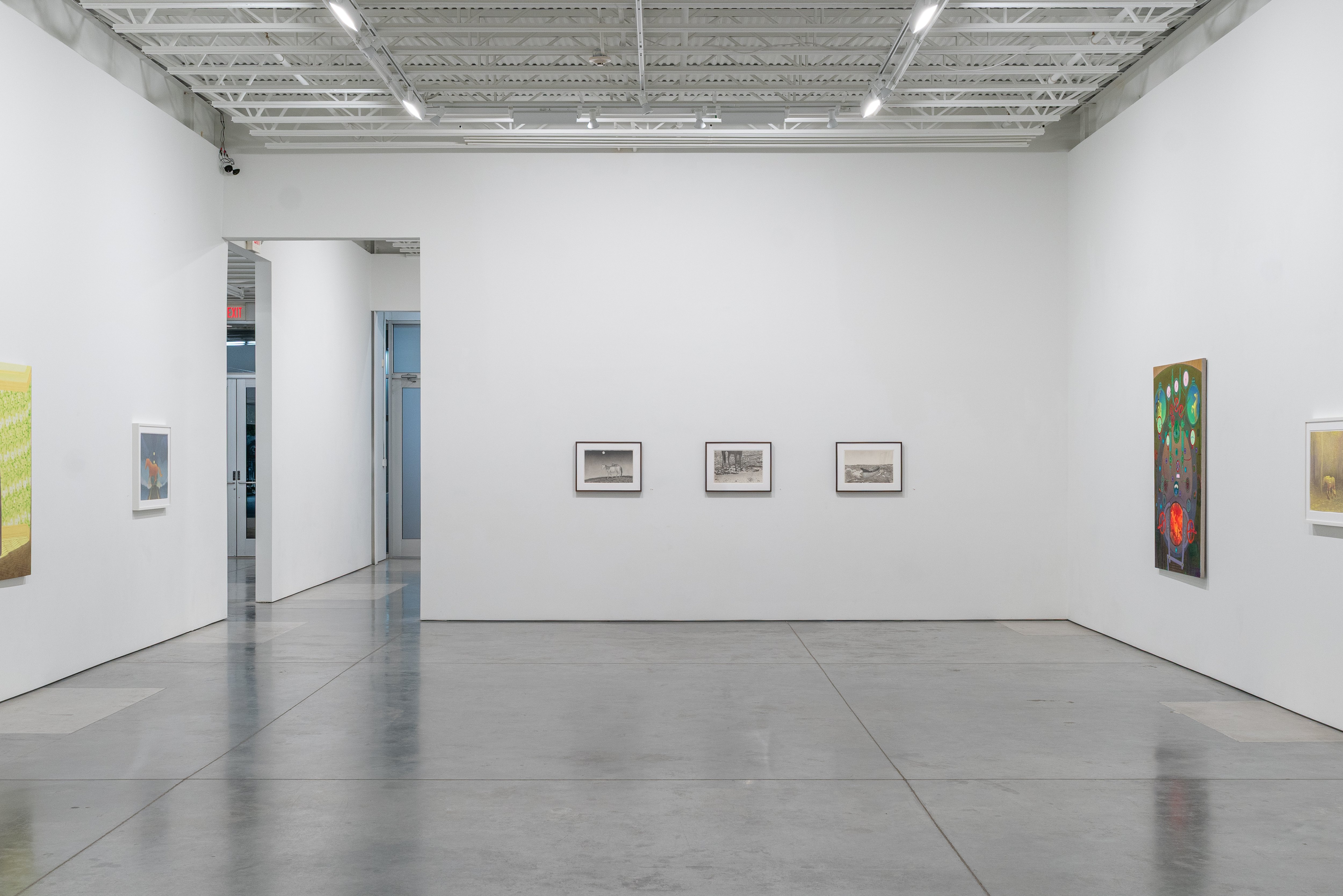 A minimalist gallery installation featuring three small framed landscape drawings hung in a horizontal line on a white wall, with larger figurative paintings on either side, set within a vast concrete-floored contemporary art space with industrial ceiling and skylights.