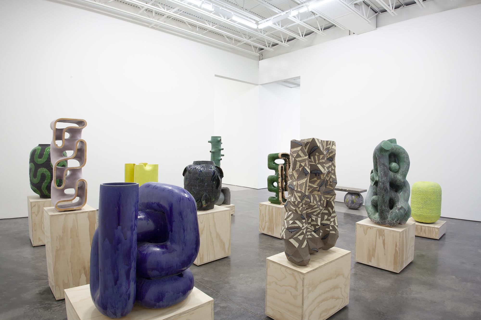 Gallery installation view featuring nine ceramic and mixed-media sculptures arranged on light wood plinths across a concrete floor. Works include hand-built clay vessels in deep blue, green, and earth tones with organic and geometric surface patterns, alongside sculptural forms in wood, cast aluminum, and concrete. Each piece is positioned individually on cubic pedestals, creating a grid-like composition beneath an industrial ceiling with exposed white metal beams.