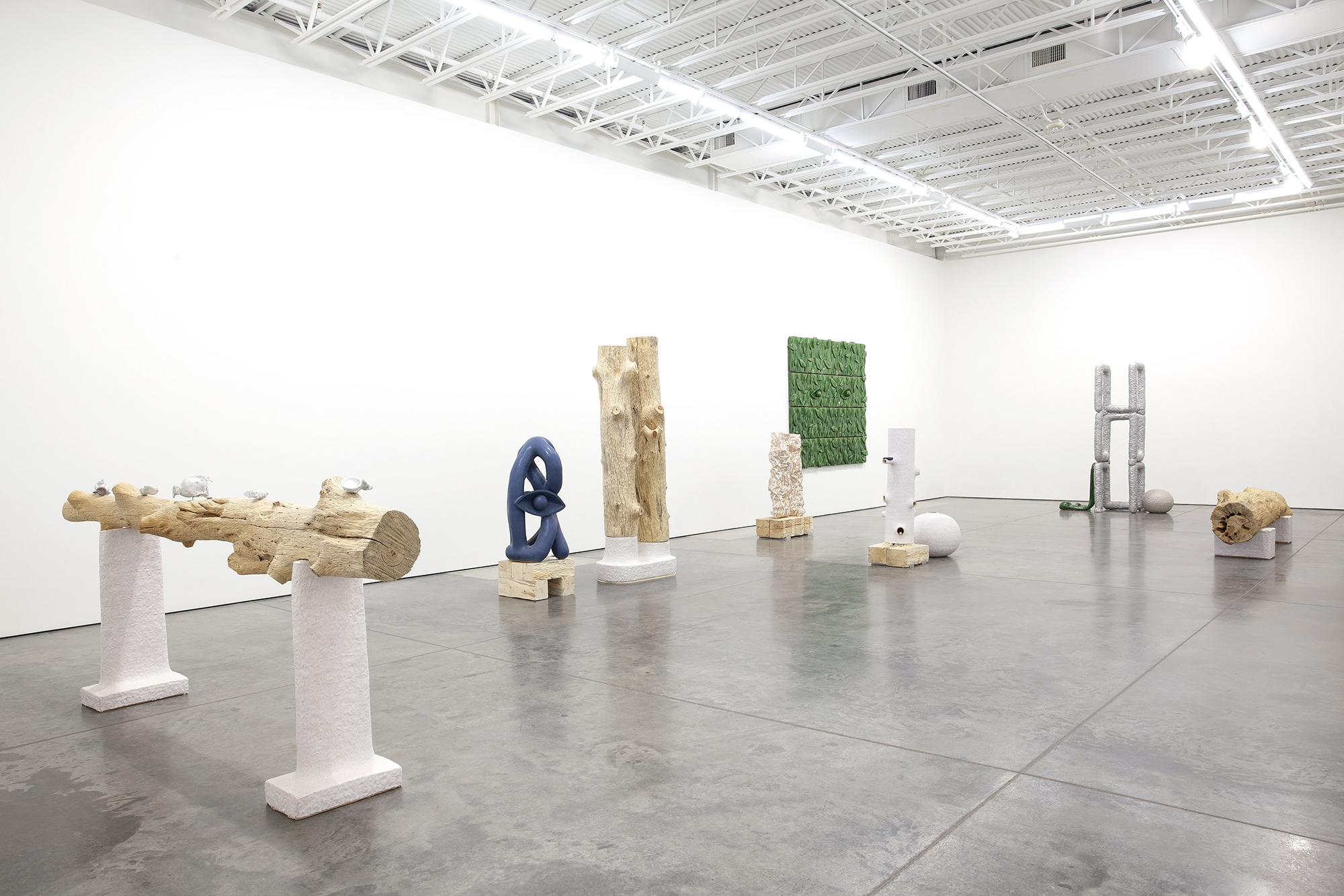 Contemporary art installation in a white gallery space featuring a linear arrangement of sculptural objects on white pedestals and plinths, including carved wooden forms, a dark blue abstract ceramic piece, natural wood columns, a green textured wall panel, cast concrete elements, and stone fragments, displayed on polished concrete flooring beneath industrial ceiling.