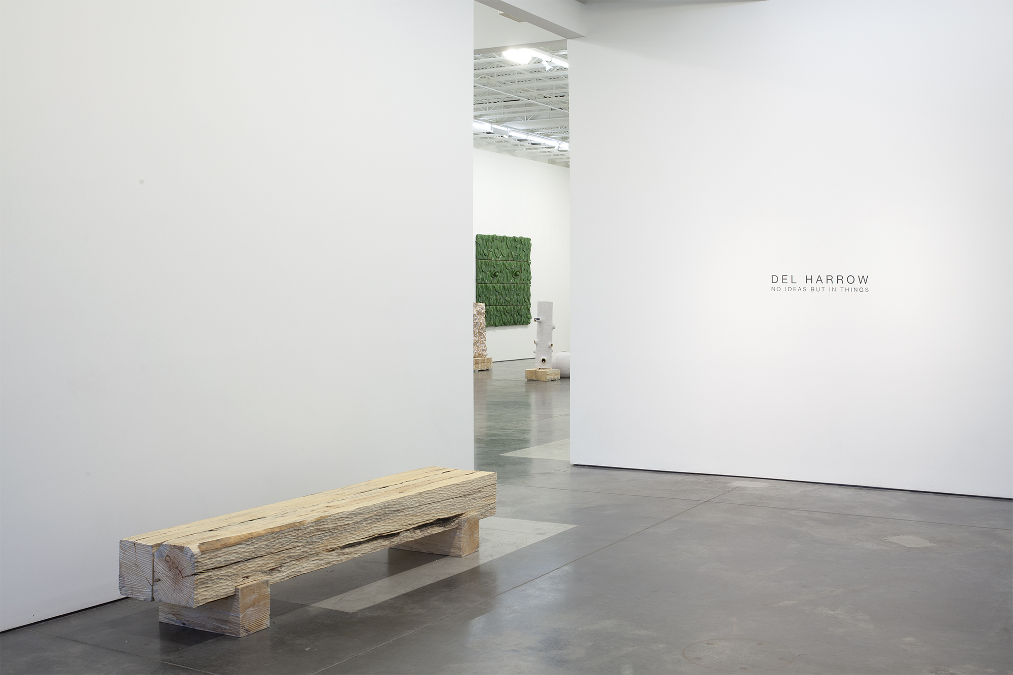 Minimalist gallery interior with white walls and polished concrete floor. A weathered wooden bench occupies the foreground. Through an open doorway, a moss-green textile panel mounted on a white wall is visible in the adjoining room, alongside wooden plinths and industrial ceiling infrastructure.